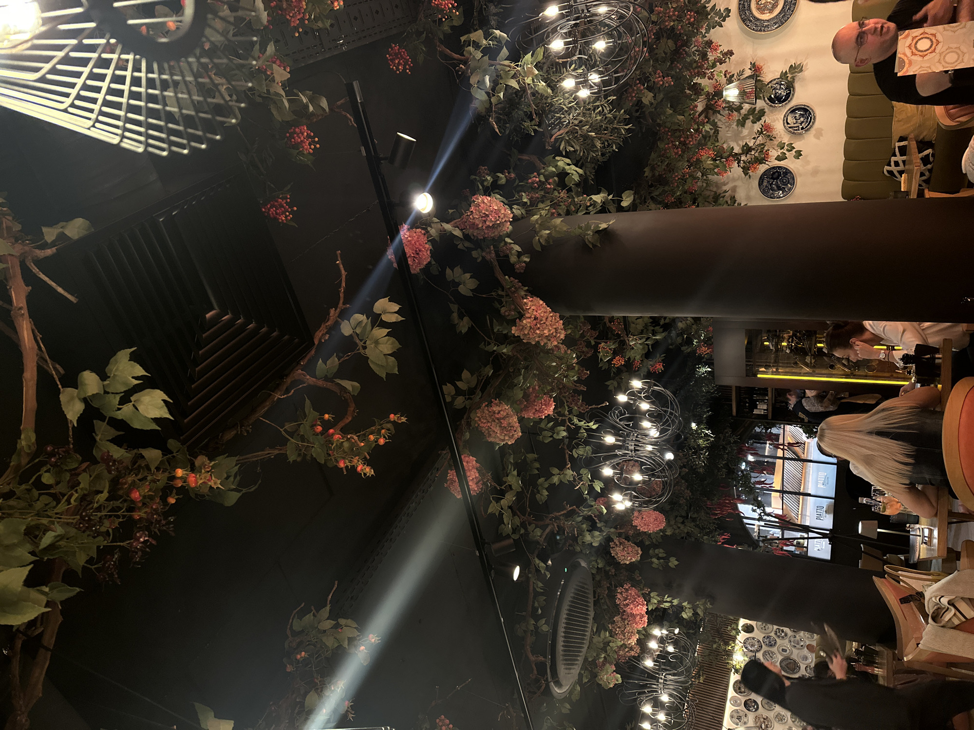 The image shows the interior of a restaurant in Sofia, Bulgaria. The ceiling is decorated with artificial flowers and light fixtures. A large, brown column runs vertically through the frame. Patrons are seated at tables, including a person with blonde hair and others along a cushioned bench. Decorative plates adorn the wall. The restaurant appears to have large windows, with the name "PIATTO" visible outside.