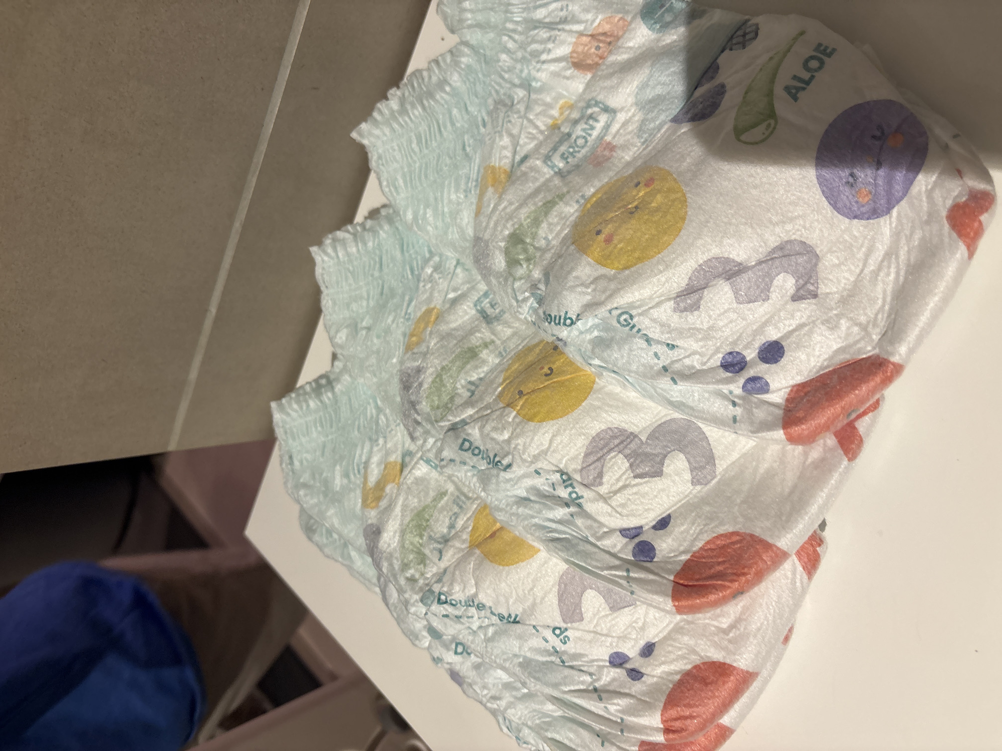 The image shows a stack of four children's diapers, size 3, arranged on a white surface. The diapers are light blue with colorful cartoon characters and graphics, including smiling faces, dots, and the word "ALOE." The word "FRONT" is visible on one of the diapers. In the background, a person wearing a blue shirt and tan pants is partially visible, along with a tan-colored wall. The scene appears to be indoors, possibly in a residential setting. There is no discernible indication of the city of Dubai or the United Arab Emirates in the image.
