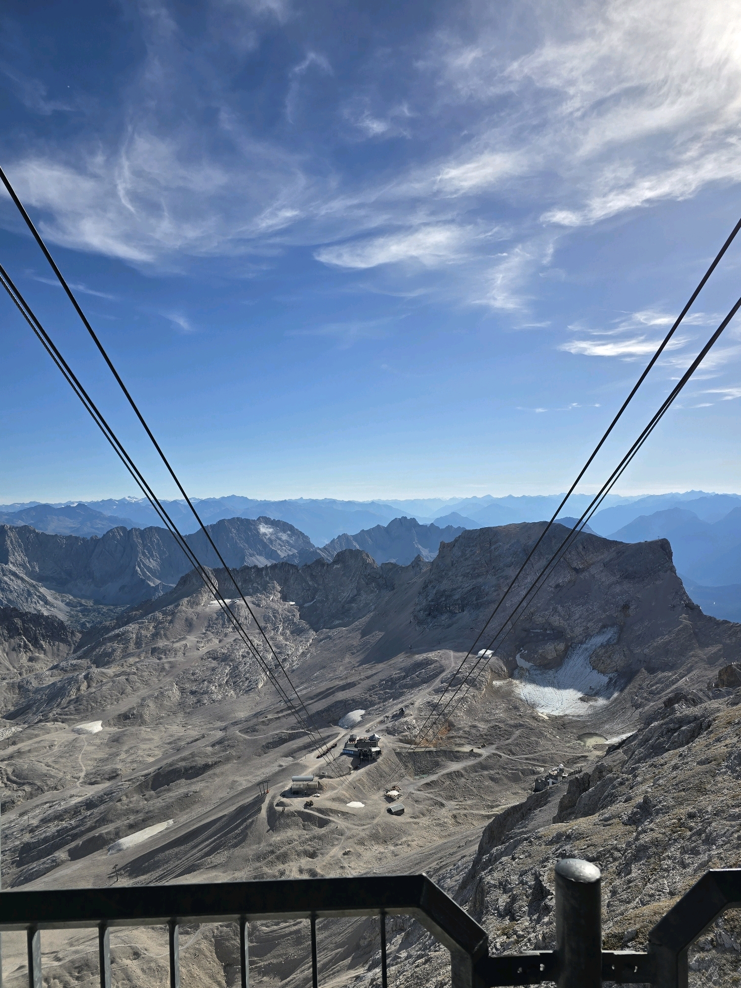 The image shows a high-angle view of the Zugspitze, Germany's highest peak, located near Grainau. The foreground features a metal railing, possibly part of a viewing platform or cable car station. Several thick cables, likely for a cable car, descend diagonally across the frame, suggesting movement or potential for transport.
The mid-ground reveals a rugged, rocky mountainous terrain. Patches of snow or ice are visible on some slopes, indicating a high altitude. A small cluster of buildings, likely part of a mountain resort or research facility, is situated on a relatively flat area within the mountains. Numerous winding paths or roads are visible across the landscape, suggesting access and visitor activity.
In the background, a vast expanse of mountain ranges stretches towards the horizon, fading into a hazy blue due to atmospheric perspective. The sky above is a vibrant blue, dotted with wispy white clouds that create a dynamic texture. The overall scene conveys a sense of scale, natural beauty, and human infrastructure in a high-alpine environment. There are no people clearly visible in the image, but the presence of buildings and paths implies human presence or recent activity.