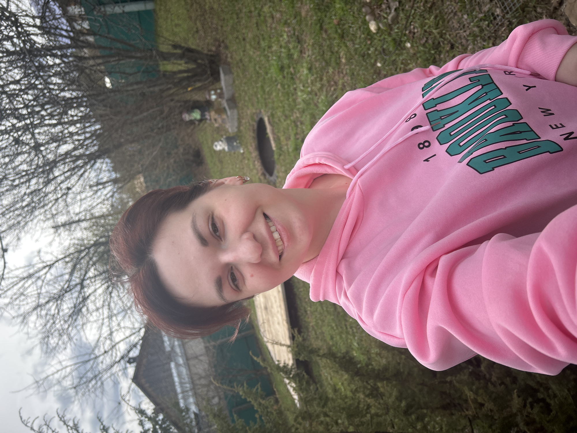 A person is pictured in a medium shot. She is wearing a pink hoodie with the text "New York 1881" printed on it. She is smiling at the camera. The background shows a yard with green grass, trees, and a fence. In the distance, there is a building and some grave markers. The photo was taken in Фоки, Russia.