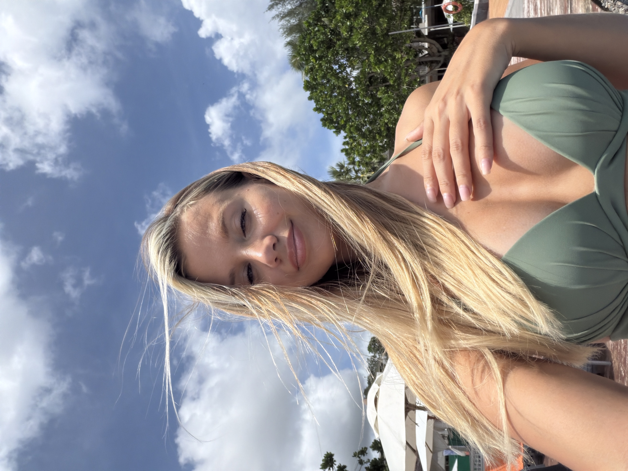 A blonde woman in a green bikini poses for a selfie outdoors, with her hand resting on her chest. Her hair is blowing in the wind. In the background, there are white umbrellas, green trees, and a cloudy blue sky. The location is Mai Khao, Thailand.