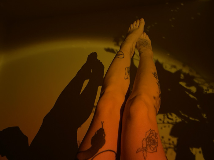 The image displays a pair of human legs, bare from the mid-thigh downwards, positioned upwards against a flat surface, likely a wall, in Boreti, Montenegro. The feet are visible at the top of the frame, slightly crossed at the ankles. The skin tone is illuminated with a warm, yellowish-orange light. Tattoos are present on both legs: an hourglass on the upper right thigh, a celestial body on the upper left thigh, a rose outline on the lower right leg, and a snake outline on the lower left leg. The background surface exhibits strong, dark shadows, including the silhouette of another pair of legs on the left side and a complex, possibly natural, pattern resembling foliage or branches on the right. The lighting appears to be artificial, casting distinct shadows and contributing to the overall warm color cast.