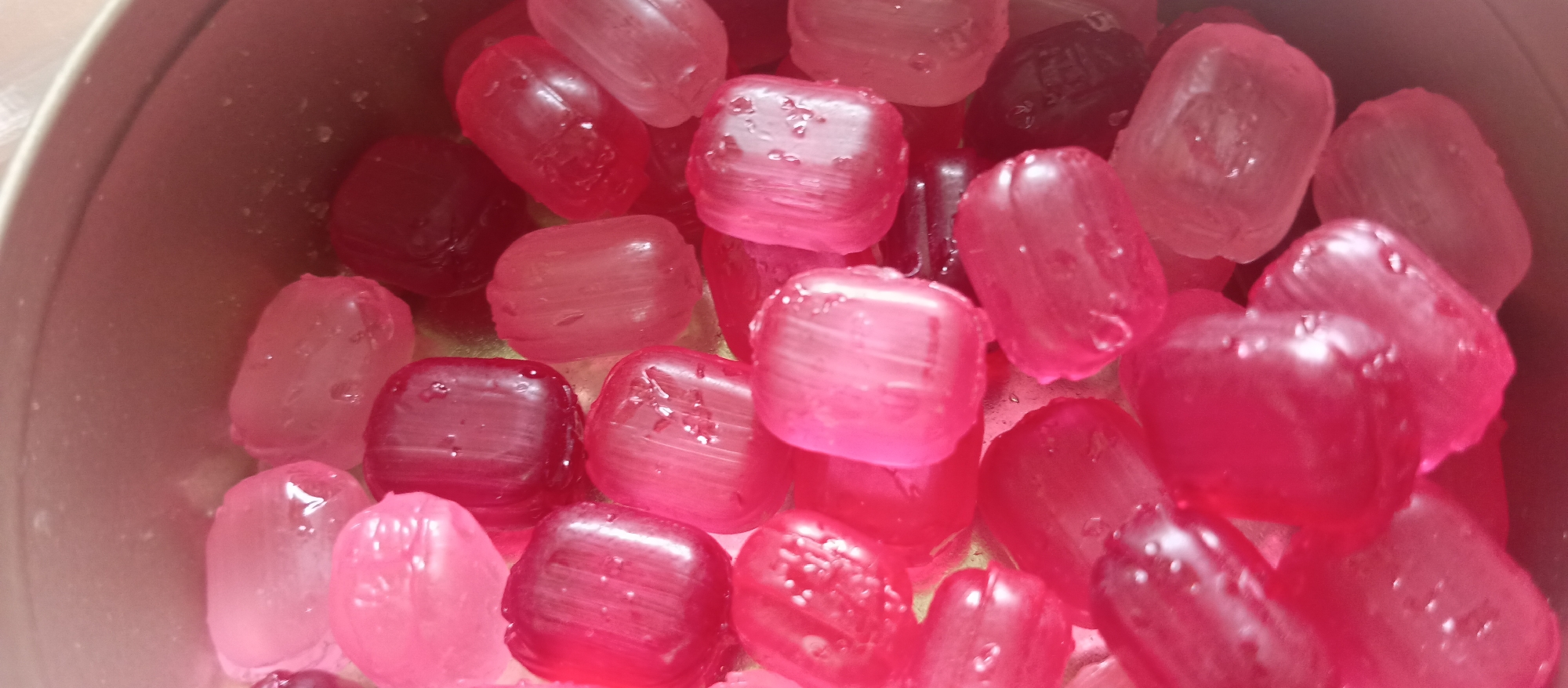 A close-up, high-angle shot displays a collection of translucent, square-shaped candies. The candies exhibit a range of red and pink hues. They are piled inside what appears to be a metal container. The candies have a slightly wet and reflective surface. The location is unidentified, as Unknown, Unknown is not a recognized city.