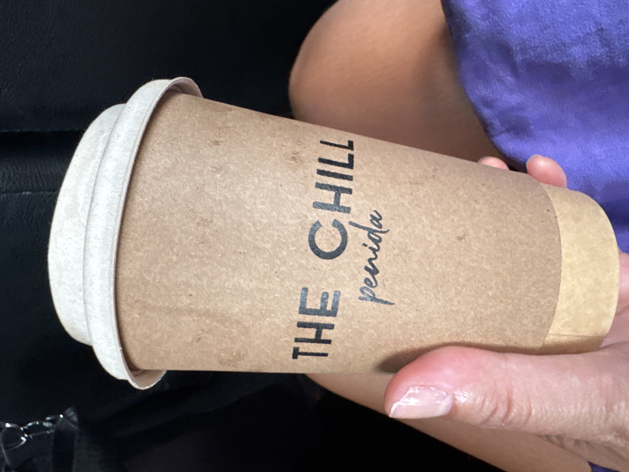 A person is holding a disposable coffee cup that says "THE CHILL penida". The cup is made of brown paper and has a white lid. The person's hand is visible, with manicured fingernails. The background is dark and out of focus, but appears to be a car interior with part of a purple garment visible. The image was taken in Nusa Penida, Indonesia.