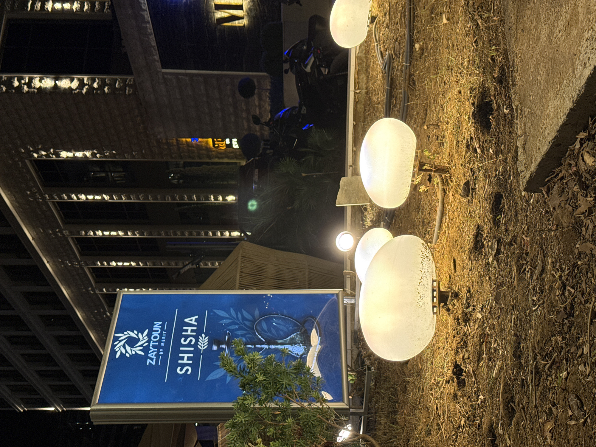 An outdoor scene at night, in the city of Budva, Montenegro, features a prominent blue sign for "ZAYTOUN BY MERIT - SHISHA" illuminated with white text and a graphic of a hookah. The sign is flanked by a small evergreen tree on the left and an outdoor seating area with woven structures in the background. In the foreground, several large, spherical, translucent lamps emit a warm, white light, casting a glow on the dry, leafy ground. Behind the lamps, a motorcycle is parked, partially obscured by foliage. The background shows the illuminated facade of a modern building with many windows.