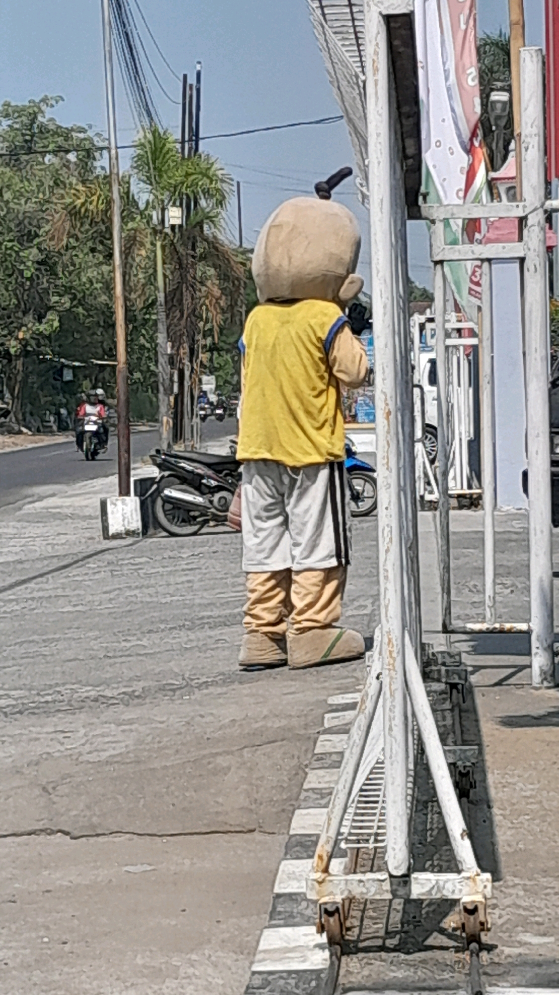 The image shows a person in a mascot costume, resembling a cartoon character with a large, round head and a small black protrusion on top. The costume is predominantly light brown for the head and legs, with a yellow shirt and light grey shorts with black and white stripes on the sides. The character is standing on a sidewalk with a black and white striped border, facing away from the camera.
In the background, a street with light traffic is visible. Several motorcycles are present, with one rider in the foreground on the left wearing a red shirt and a white helmet. Trees and utility poles with numerous electrical wires are also visible, suggesting an urban or suburban environment. The sky is clear and blue.
On the right side of the image, a white metal structure with a slatted design is prominent, partially obscuring the view of the street and background. Several vertical banners with various colors and designs are attached to the structure, some of which appear to have writing on them.
The overall scene is outdoors, during daylight, with a moderately busy street in the background. The location is identified as Tanjung, Indonesia, based on the context provided.