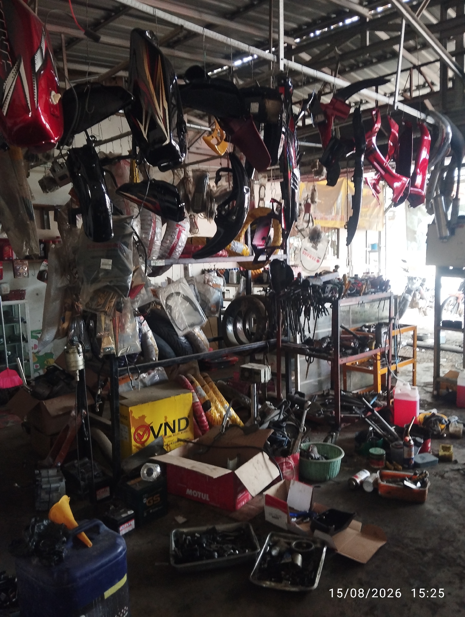 The image depicts an interior scene of what appears to be a motorcycle repair shop or parts storage facility in Tegal Rejo, Indonesia. The space is cluttered with various motorcycle parts, tools, and equipment. Motorcycle body panels, such as fuel tanks and fenders, are predominantly red and black, some with decorative graphics, and are hung from the ceiling on hooks. Shelving units are laden with more parts, tires, and boxes. In the foreground, a blue plastic container with a yellow funnel is visible, alongside other containers and tools scattered on the floor. A yellow box with the text "VND" and a red circular logo is prominent on a shelf. Several batteries, including one with a "GS" logo, are also present. Various tools, including wrenches and pliers, are organized on metal racks. The overall environment suggests a working garage with items in storage and potentially undergoing repair or assembly. The lighting is a mix of natural light filtering in from outside and artificial lighting from the ceiling.