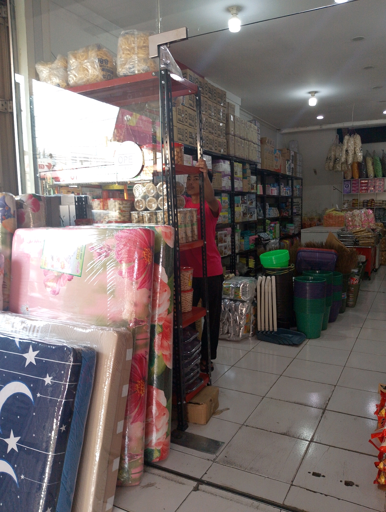 The image depicts an indoor scene within a store located in Lembang, Indonesia. The store appears to be a general goods or household item retailer. In the foreground, several rolled mattresses are stacked and wrapped in plastic. One mattress features a blue pattern with white stars and moons, while others have floral designs.
The store's interior is lined with tall shelving units filled with various products. On the left side, there are shelves stocked with packaged goods and what appear to be food items in clear bags. A large white sign with the word "OPEN" in a circle is visible above the shelves.
A person wearing a red shirt and black pants stands near the shelving units, appearing to be an employee or shopper. The person is holding onto a metal shelf pole and is facing towards the right side of the image.
The shelving units on the right display a wider assortment of items, including boxes, containers, and possibly cleaning supplies or toiletries. On the floor in the middle section of the image, there are stacks of plastic buckets and containers in various colors, including green and purple. There are also brooms leaning against the shelves and a shallow blue plastic tray.
The overall environment suggests a retail space with a variety of merchandise arranged for sale. The lighting in the store appears to be a mix of overhead fluorescent lights and possibly natural light from the entrance. The floor is tiled.