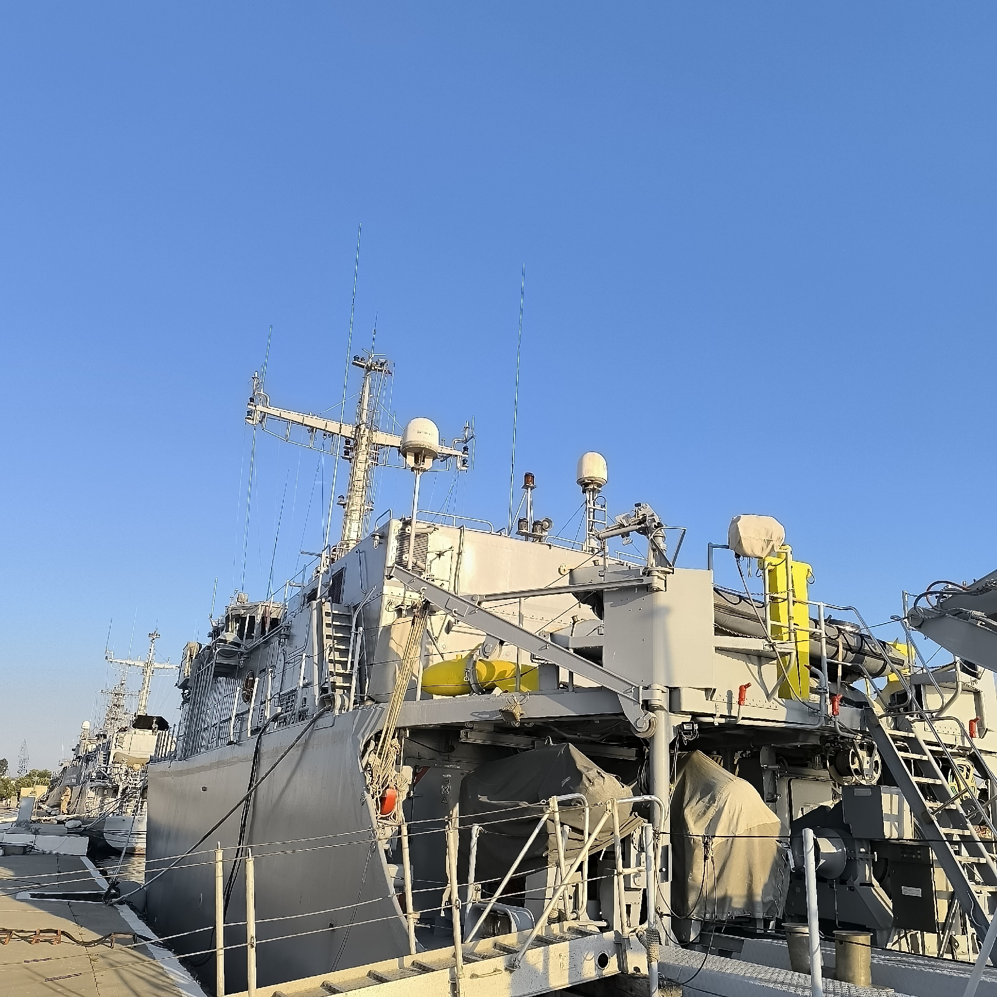The image shows a line of naval vessels docked at a pier under a clear blue sky. The ships are gray and appear to be modern military vessels, with various antennas, radar domes, and equipment visible on their decks. The closest vessel is in sharp focus, revealing details like ladders, railings, and covered equipment. In the background, other ships are visible, as are parts of a city's skyline and greenery in the distance, suggesting an urban port environment. The scene is static, with no people or activity depicted. The lighting suggests it is daytime, likely morning or late afternoon due to the angle of the light. This is a view of a naval port in Varna, Bulgaria.