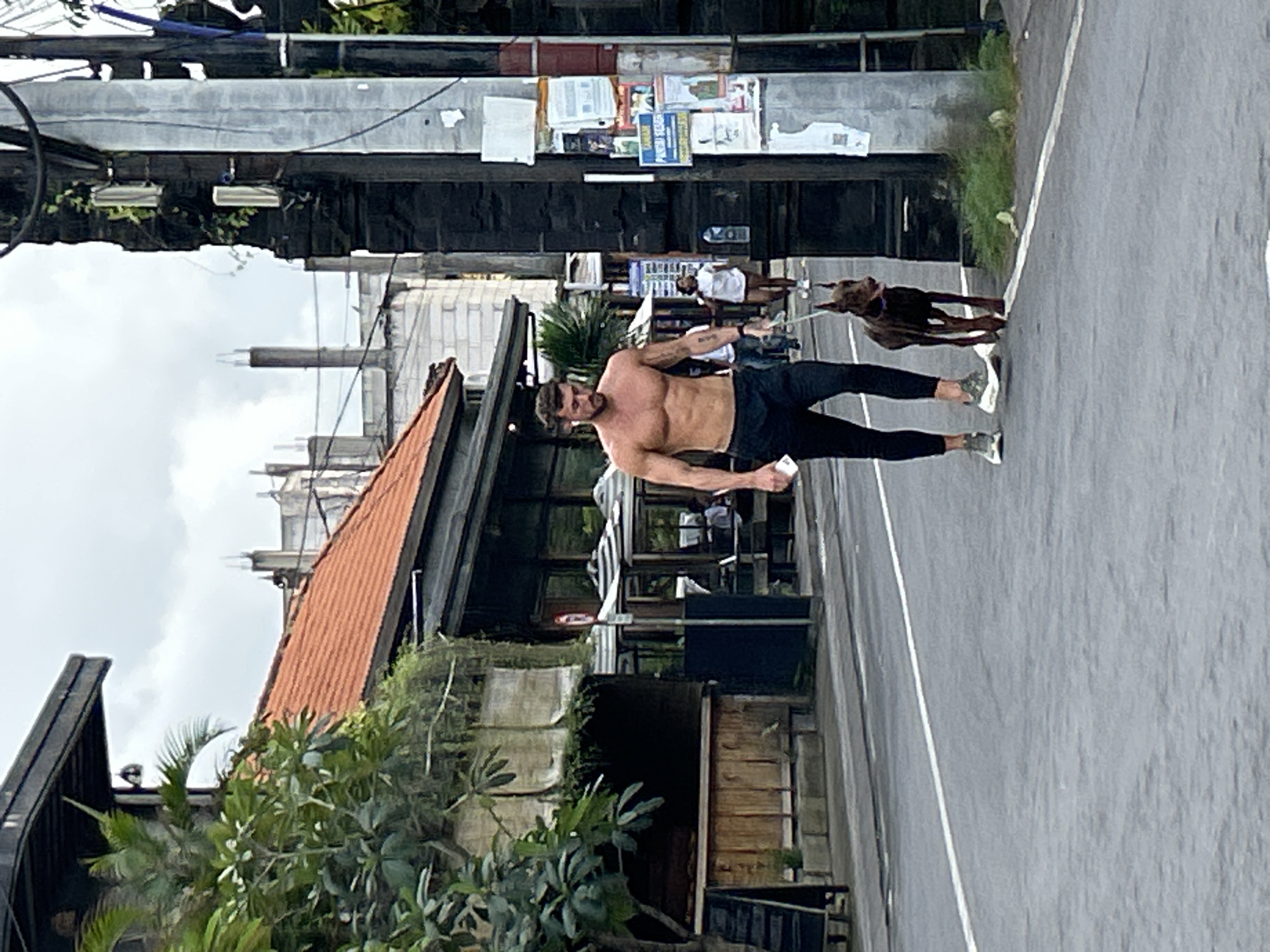 A man is walking a dog on a street in Mengwi, Indonesia. The man is shirtless and wearing black pants and sneakers. The dog is a dark brown Doberman Pinscher. In the background, there are buildings with red tile roofs, trees, and power lines. The sky is cloudy. The scene appears to be outdoors on a paved road.