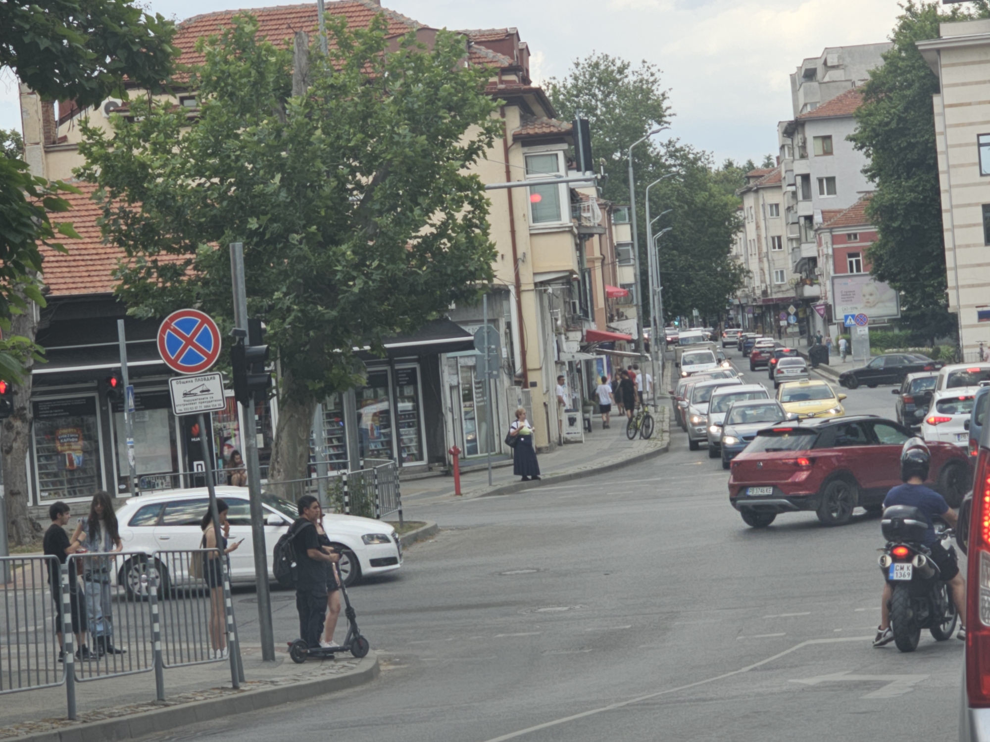 The image depicts a street scene in Plovdiv, Bulgaria, likely during the daytime under an overcast sky. The foreground shows a paved road with traffic, including cars, a motorcycle, and a scooter. On the right side of the road, a man is riding a motorcycle. In the middle ground, there are several buildings lining the street, some with businesses on the ground floor. A white station wagon is parked on the left side of the road. Several people are visible on the sidewalk, some standing and others on a scooter. Traffic signs, including a red and blue circular sign with a diagonal line through it, are present. The background shows more buildings and trees. The architecture appears to be a mix of older and more modern structures.