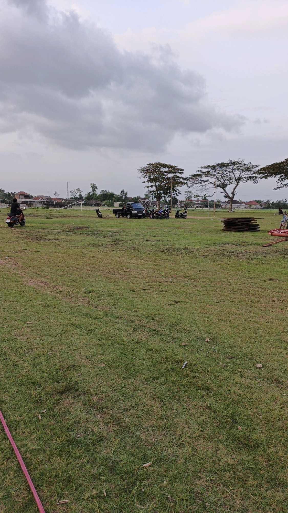 An open, grassy field is pictured under a cloudy sky. The field is mostly flat with some patches of dried grass. In the background, a line of houses with red roofs stretches across the horizon, suggesting a residential area. Several trees, some with broad canopies, are scattered throughout the scene. A black pickup truck is parked in the middle ground, surrounded by several motorcycles. A person is riding a motorcycle on the left side of the image. To the right, a stack of dark, rectangular objects, possibly wood or building materials, is visible, along with some red construction piping. The location is identified as Boyolangu, Indonesia.