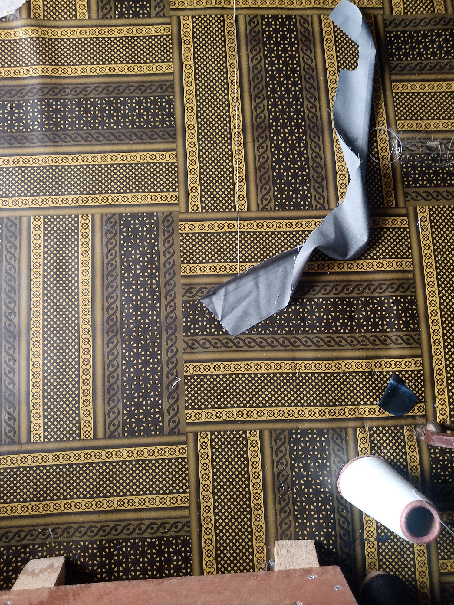 An overhead shot depicts a tiled floor in Jauro Yinu, Nigeria, covered with a patterned fabric. The fabric features a repeating design of golden yellow and dark brown geometric shapes. A section of light gray fabric is draped across the patterned floor, with its edges curling upward. In the bottom right corner, a spool of white thread is visible, along with a small dark blue fabric scrap and part of a wooden object.