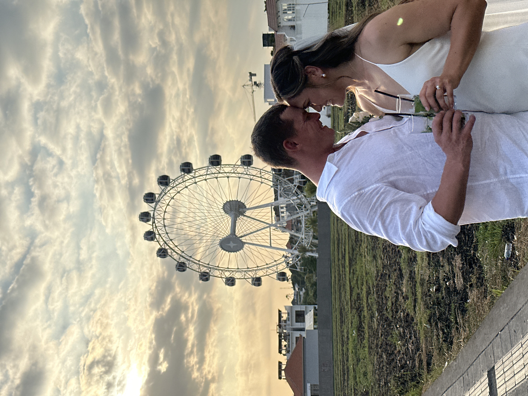 In North Kuta, Indonesia, at sunset, a newlywed couple stands close, foreheads touching, with a Ferris wheel visible in the background. The groom, wearing a white linen shirt, holds two glasses of mojitos, one of which he offers to the bride. The bride, in a white wedding dress, holds the other glass, and her engagement ring is visible on her left hand. The couple is positioned in front of a field with rows of dark soil and scattered green grass. In the mid-ground, there are buildings with red roofs and white facades, and further back, a construction crane is visible. The sky is filled with dramatic clouds, catching the warm hues of the setting sun. The foreground features a tiled pathway.