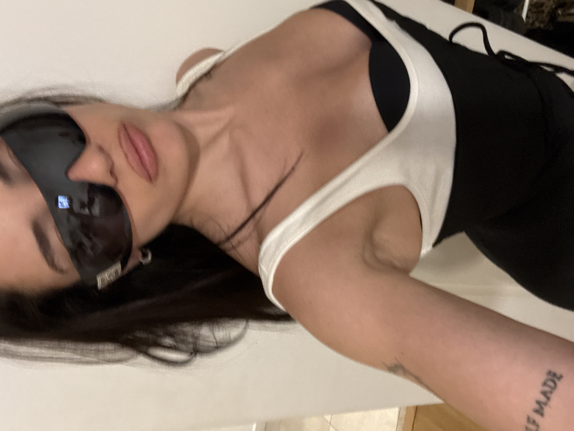 A woman is photographed in a close-up shot, angled. She wears sunglasses and a black and white outfit. Her arm is visible with a tattoo that reads "SELF MADE." The environment is unclear, but the background appears to be a white surface. The location is not discernible, and there is no indication of Buenos Aires, Argentina.