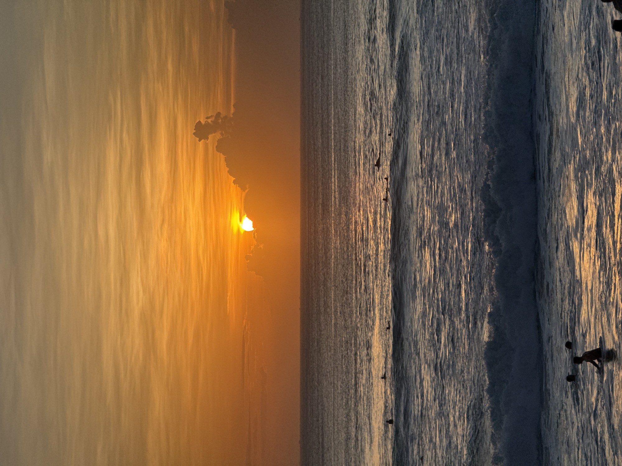The image captures a horizontal view of a sunset over the ocean. The left portion of the image features the sky with the setting sun partially obscured by clouds, casting an orange glow across the scene. The right portion of the image depicts the ocean with visible waves. Several people are scattered within the waves. The image is taken in Unknown, Unknown.