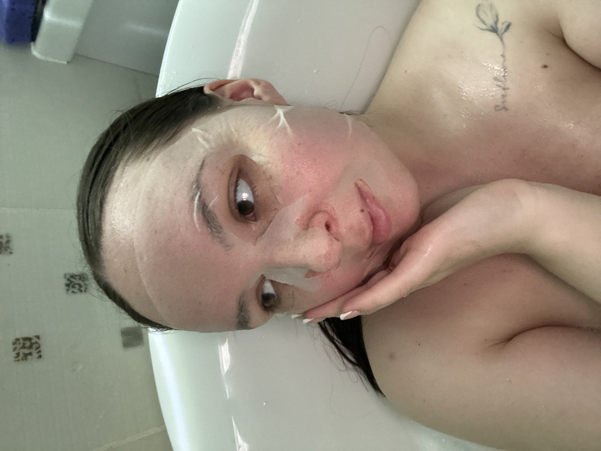 A woman is lying in a bathtub, wearing a facial mask. Her head is tilted to the side, and she is looking at the camera. Her right hand is resting on her chin. She has a tattoo on her upper chest. The background shows a white tub and tiled walls. The location is in a bathroom; the city of Chaikovsky, Russia, is not deducible from the image.