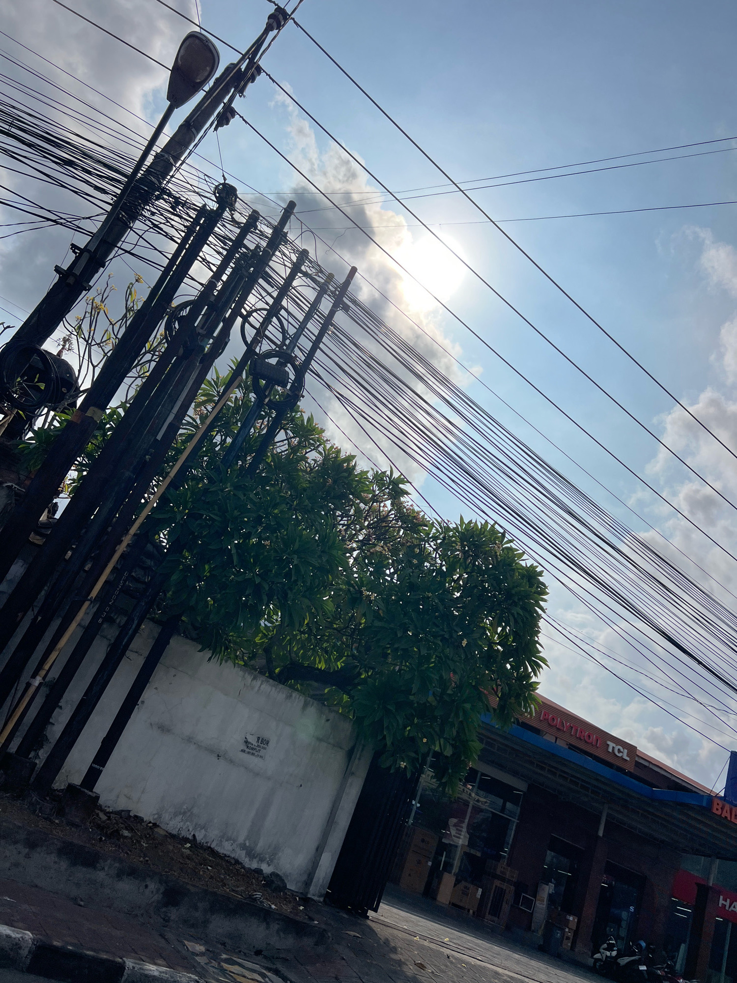 The image depicts a street scene in Denpasar, Indonesia, under a bright sky with scattered clouds. Prominently featured is a dense network of overhead electrical and telecommunication wires, supported by a utility pole with a street lamp. A lush green tree with white flowers grows near a white concrete wall. In the background, a building with a prominent sign reading "POLYTRON TCL" is visible, suggesting it is an electronics store. Several motorcycles are parked in front of the store. The overall impression is a typical urban streetscape, capturing the interplay of infrastructure, nature, and commerce.