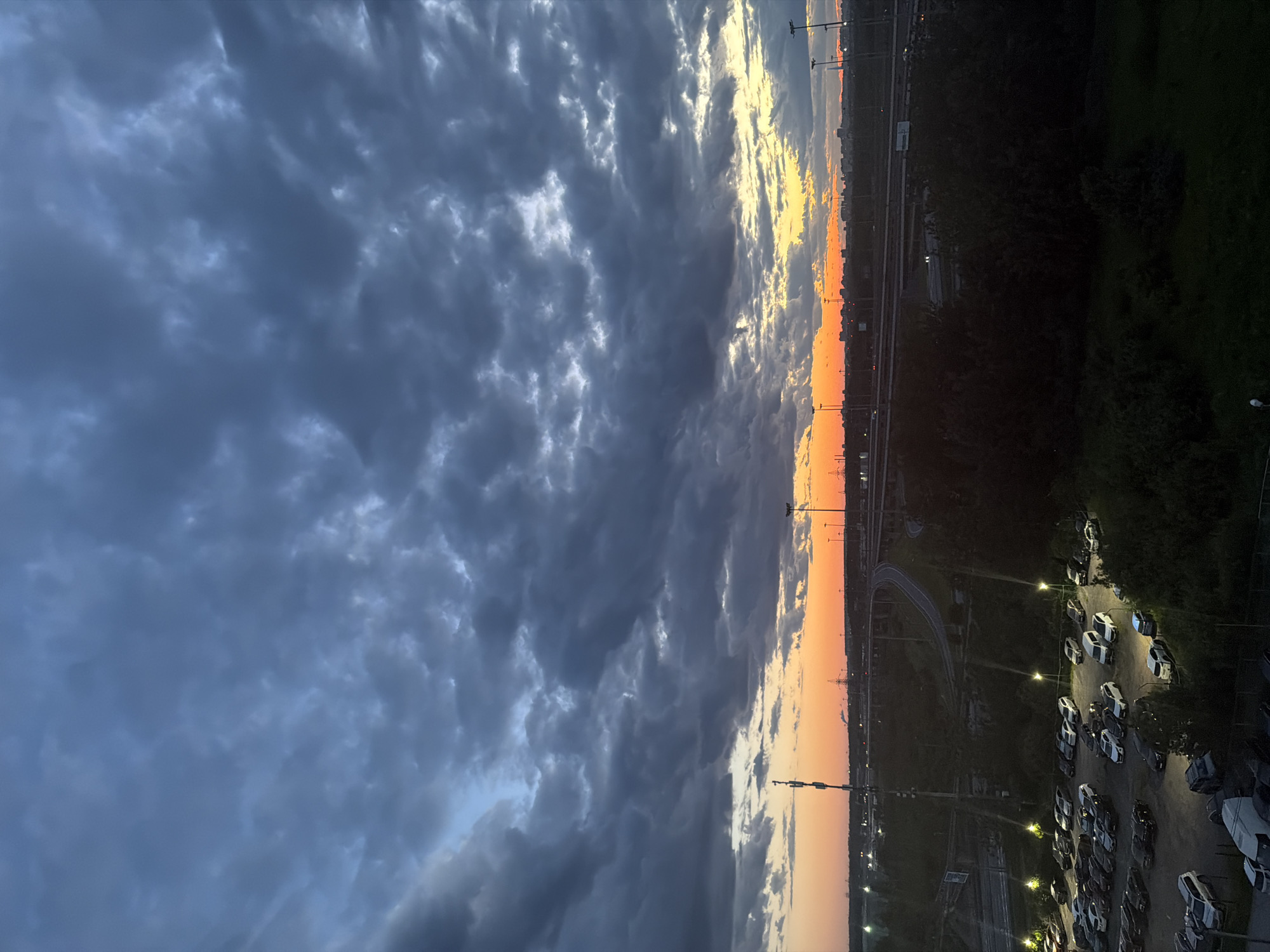 An eye-level, medium shot captures a vast expanse of dark, textured clouds that dominate the upper portion of the frame. Below, a dramatic sunset casts an orange and yellow glow on the horizon, illuminating the edges of the clouds with a warm light. The scene transitions to a dusky landscape featuring a multi-lane highway with overhead lighting, curving through dark, tree-covered terrain. A dimly lit parking lot filled with numerous cars, arranged in rows under the glow of streetlights, is visible in the foreground. A tall communication tower stands prominently on the left side of the image, with a distant cityscape silhouetted against the vibrant sunset. The overall atmosphere is one of twilight in an urban or suburban environment, marked by both natural light and artificial illumination.