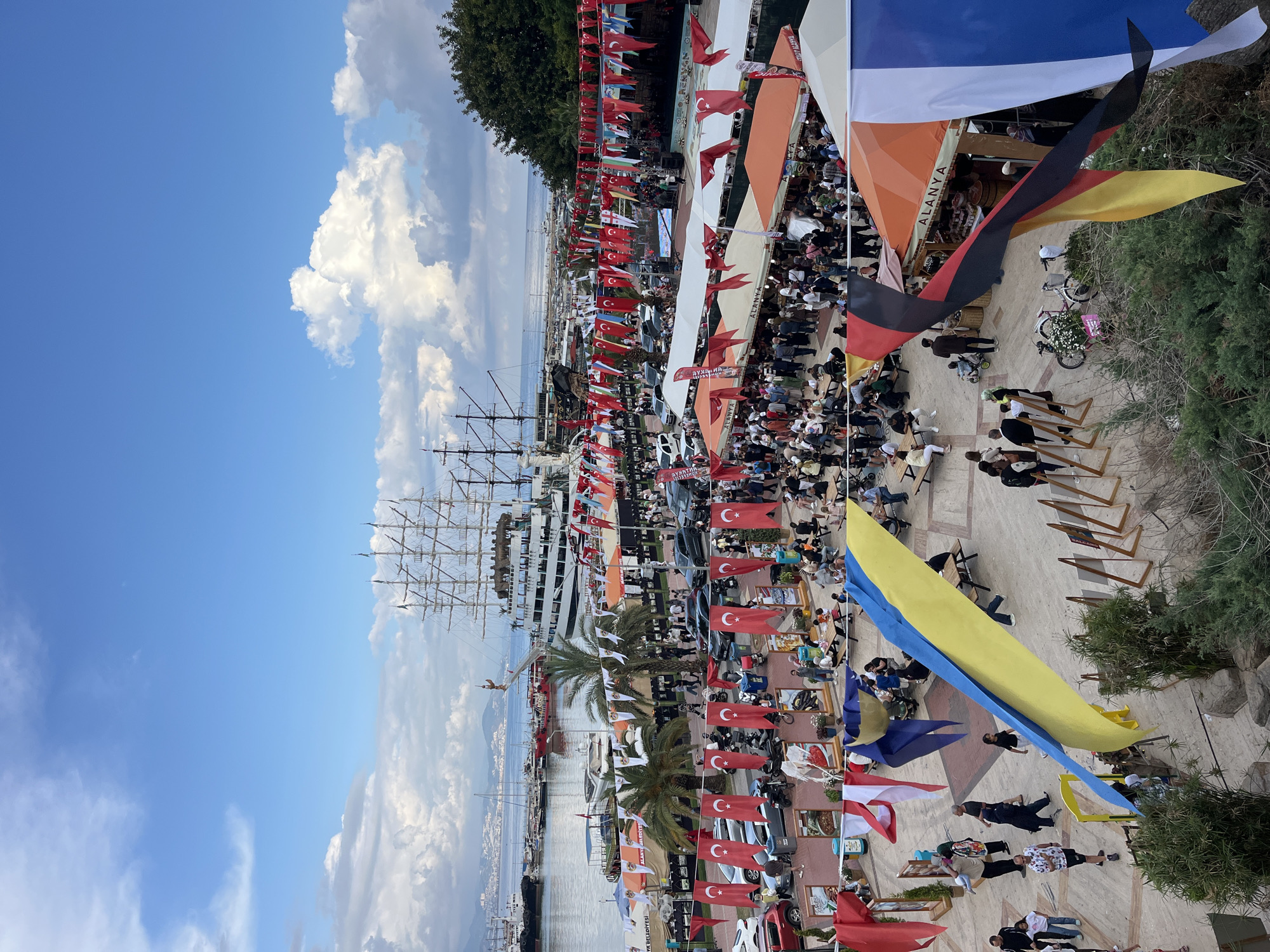 The image shows an aerial view of a crowded outdoor event in Alanya, Turkey. A large number of people are walking and gathering along a paved area decorated with numerous national flags, predominantly Turkish flags. Several stalls and tents are set up, suggesting a market or festival atmosphere. A tall sailing ship is docked nearby, visible on the left side of the frame. The scene is set against a backdrop of the sea, with a cloudy blue sky above. Trees and greenery are present on the right side of the image. The overall impression is that of a lively public gathering.