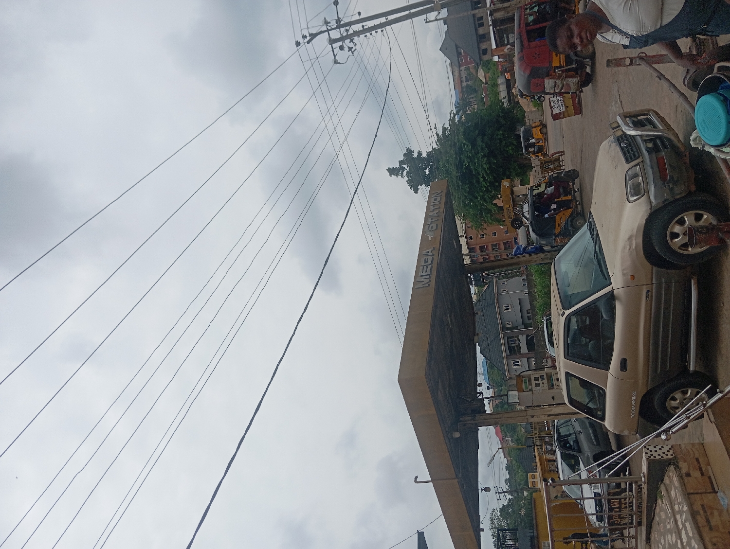 The image depicts a bustling street scene in Nkpor, Nigeria, with a gas station, vehicles, and a man, under an overcast sky during the daytime.
In the foreground and to the right, a cream-colored Toyota RAV4 SUV is parked, with its passenger side facing the viewer. The text "TOYOTA RAV4" is visible on its side. To the far right, a man in a dark shirt and light trousers is standing near the back of the SUV, looking towards the left side of the frame, possibly attending to the vehicle or conversing. A blue container is visible near him, on the ground next to the vehicle.
Dominating the midground is a gas station canopy, which spans across the image from right to left. The words "MEGA - STATION" are clearly written on the side of the canopy. Below the canopy, parts of gas pumps are visible, and other vehicles, including a dark-colored car, are parked or waiting.
Beyond the gas station, several tricycles, locally known as Keke Napep, are visible. One red tricycle is prominent, and several yellow ones can be seen further back. Some of these tricycles appear to be waiting or driving on a road. In the background, various multi-story residential or commercial buildings are scattered amongst green trees and vegetation, indicating an urban or semi-urban setting.
Above the entire scene, numerous power lines stretch across the cloudy, grey sky, connected to utility poles visible on the left side of the image. The weather appears to be overcast or cloudy, contributing to a muted lighting. The overall impression is one of daily activity in a lively community.