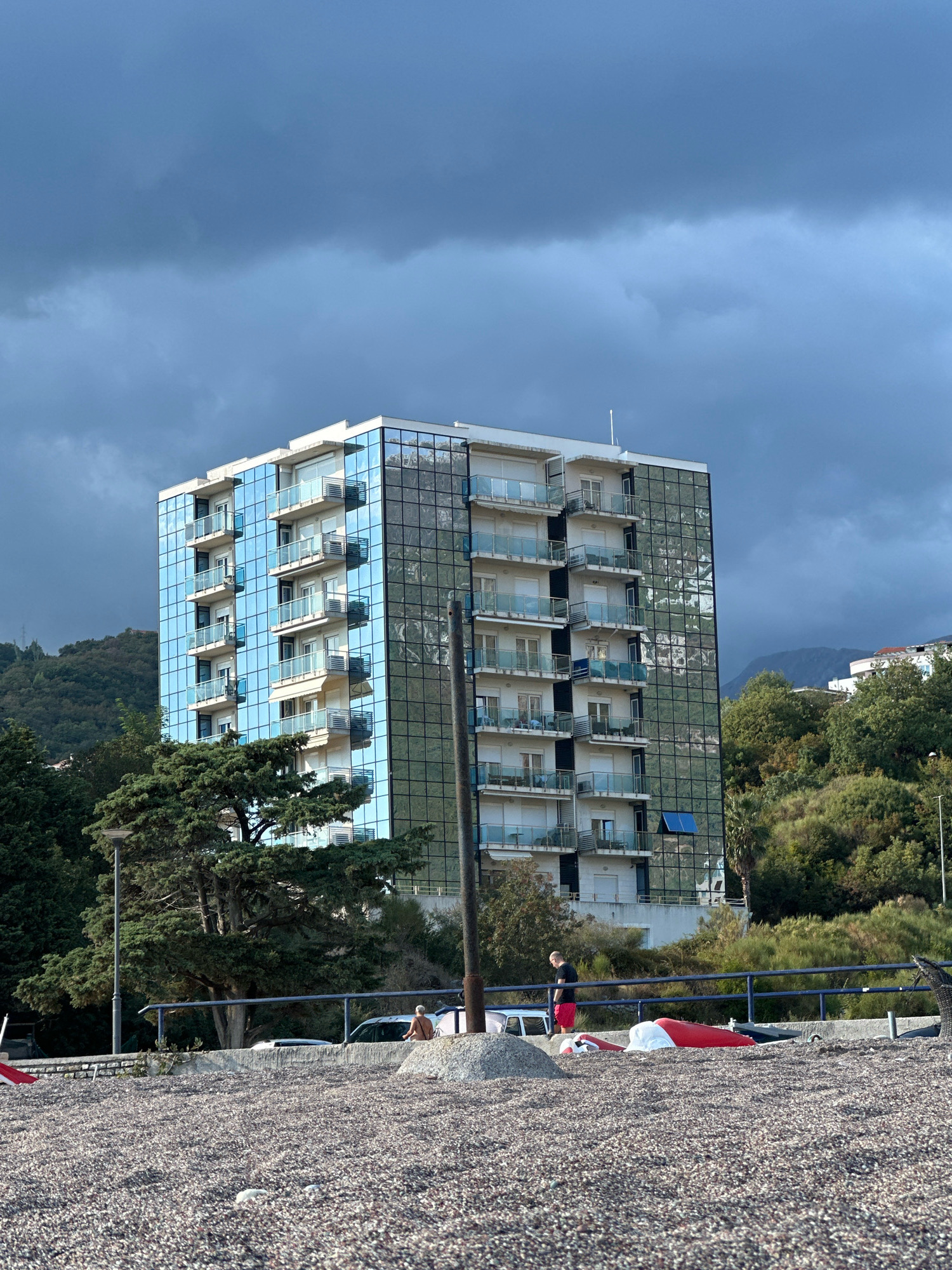 A seven-story modern building with a facade of reflective blue glass and white balconies stands against a dark, cloudy sky. The building is situated in Boreti, Montenegro, surrounded by lush green trees and vegetation on a hillside. In the foreground, a pebbled beach is visible with a blue railing separating it from a road. Several objects, including a red and white inflatable, a car, and a person in red shorts and a black shirt, are present on the beach and near the railing. A lamppost stands to the left of the building, and a dark pole with cables is to its right. The overall scene suggests an overcast day near the coast.