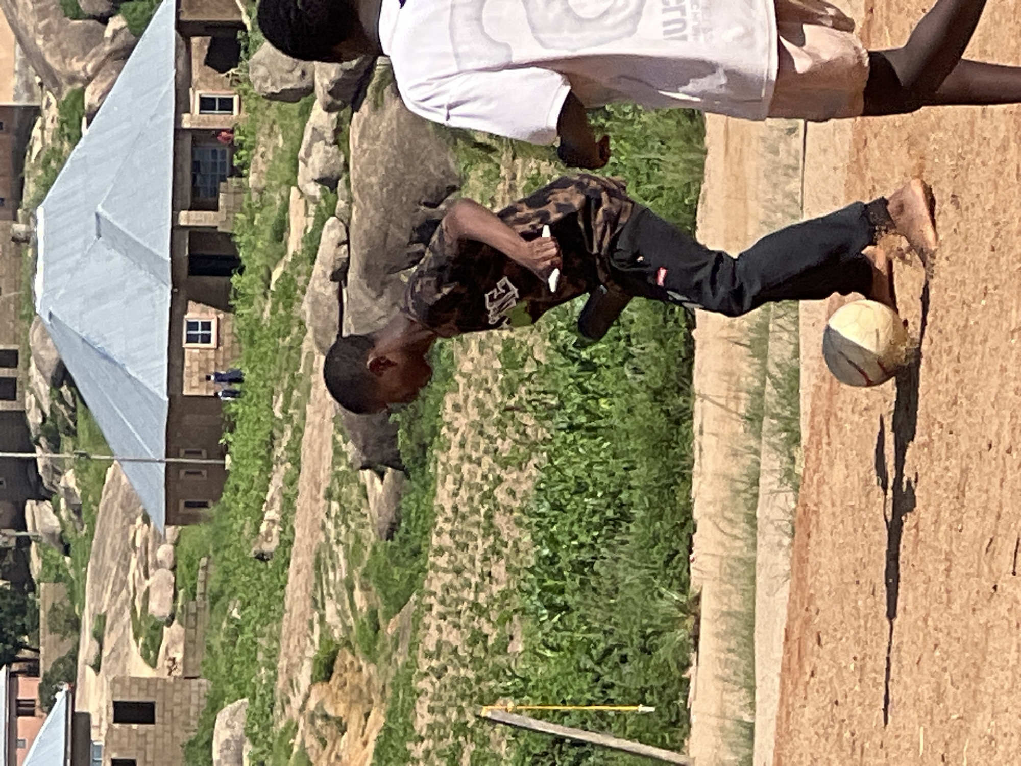In the city of Jos, Nigeria, a young boy in a patterned t-shirt and black pants is captured mid-stride, his bare feet kicking a soccer ball on a dusty, reddish-brown ground. He is looking towards the ball, his body angled as if about to make a play. Behind him, another individual, partially visible from the back, wears a white t-shirt and light-colored shorts. The background reveals a rural landscape with sparse vegetation and numerous large rocks scattered across rolling hills. In the distance, several buildings with corrugated metal roofs are visible, suggesting a residential area. A yellow rope hangs from a pole on the left side of the frame, adding a touch of everyday life to the scene. The lighting indicates it is daytime, with shadows cast on the ground, suggesting a sunny environment.