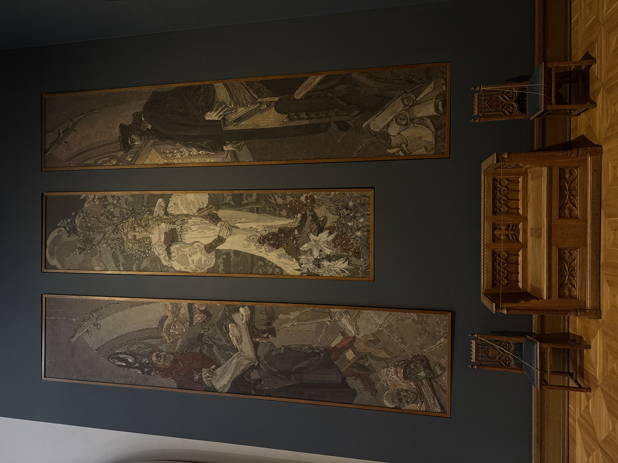 The image shows a wall in a room, with three large, horizontally oriented framed artworks hanging on it. The artworks appear to be paintings or murals with detailed figures. Above and to the right of the artworks, there is a wooden structure with carved details, possibly a cabinet or part of a larger architectural element. Two small wooden chairs with blue seats are attached to the side of the wooden structure.
The wall is painted in a dark blue-gray color, and the wooden structure and chairs are made of a lighter-colored wood. The floor appears to be wood with a parquet pattern. The image was taken in the Tretyakov Gallery in Moscow, Russia.