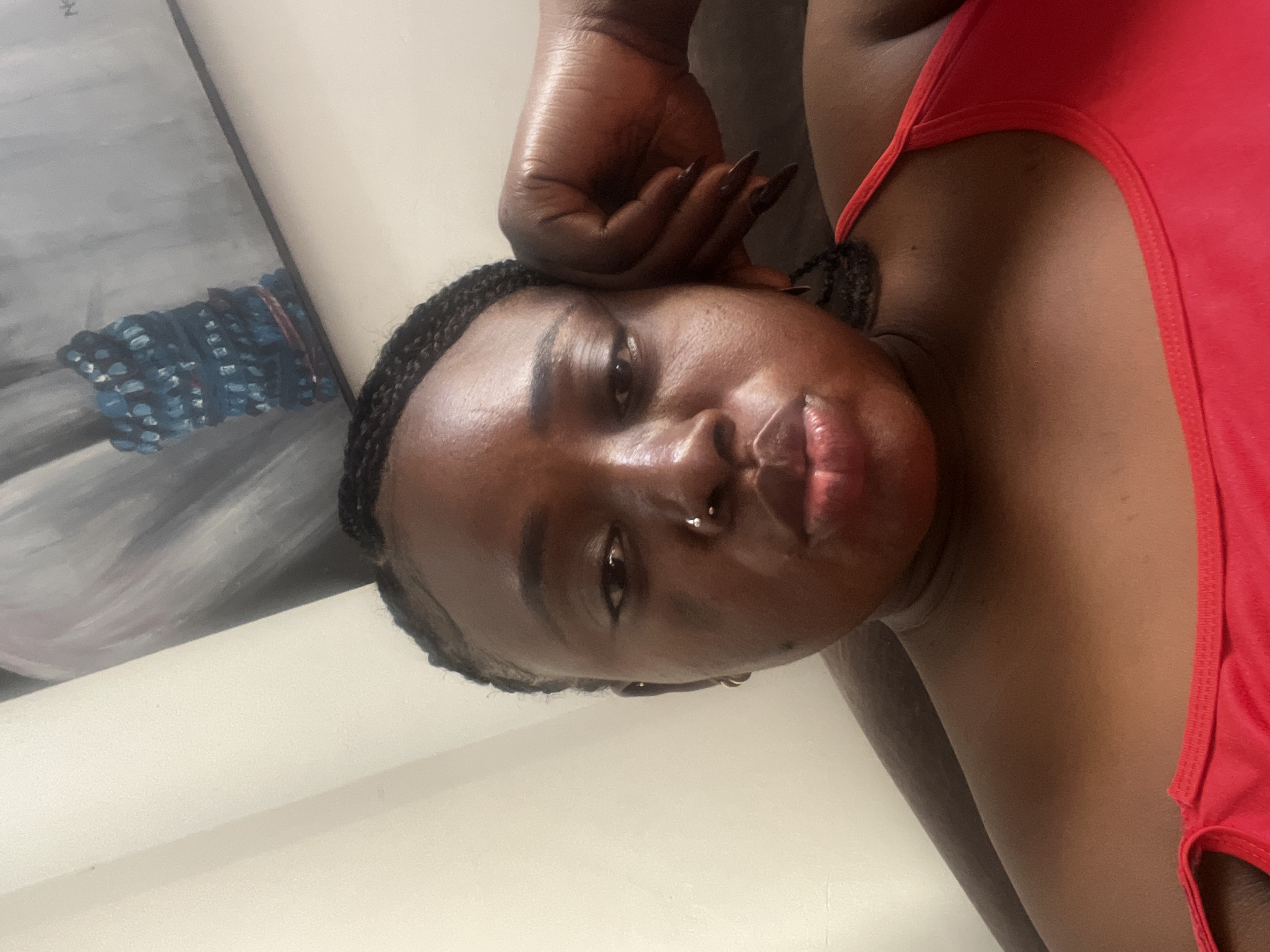 A medium shot shows a Black woman with braided hair, wearing a red tank top and a nose ring, resting her head on her hand. The background features a painting and a plain wall. The woman is positioned in the center of the frame, looking towards the camera with her lips slightly pursed. Her right arm is bent, with her hand supporting her head. The lighting is soft, illuminating her face and upper body. The painting in the background appears abstract, with muted colors and a focus on a textured blue element. The overall setting suggests an indoor environment, possibly a home or studio in Abuja, Nigeria, given the context provided.