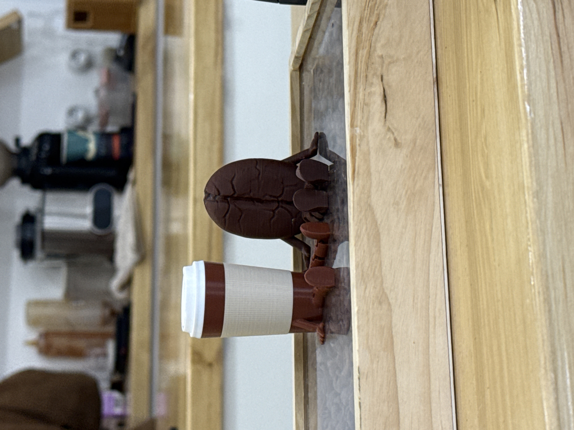 A 3D-printed coffee bean figurine, holding a disposable coffee cup with a white lid and a cream-colored middle section, stands on a ledge. The figurine and cup are light brown. The ledge appears to be part of a light-colored wooden structure. In the blurred background, other objects are visible, including a coffee grinder and various bottles and containers. There is no verifiable information to place this image in Krasnoyarsk, Russia.