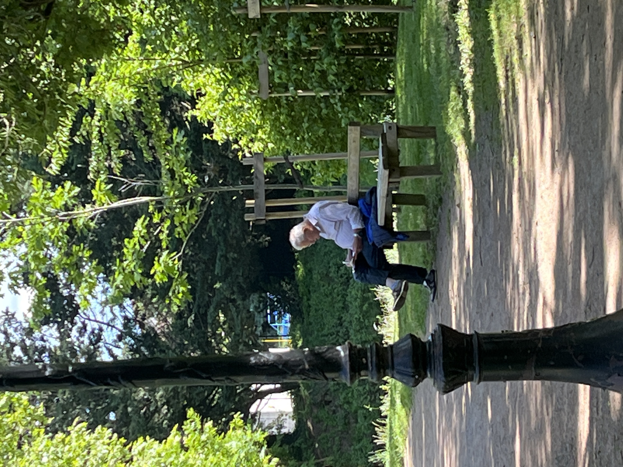 The photograph depicts a scene in what appears to be a park in Saint-Galmier, France. An older man is seated on a wooden bench, leaning forward. He is wearing a white shirt, dark pants, and what seem to be sneakers. A blue jacket is draped over the back of the bench next to him.
The environment includes lush greenery and trees, creating a natural backdrop. The bench sits beside a dirt or gravel path, with dappled sunlight creating shadows along the ground. A black, ornate lamppost is prominently featured in the foreground, partially obscuring the view.