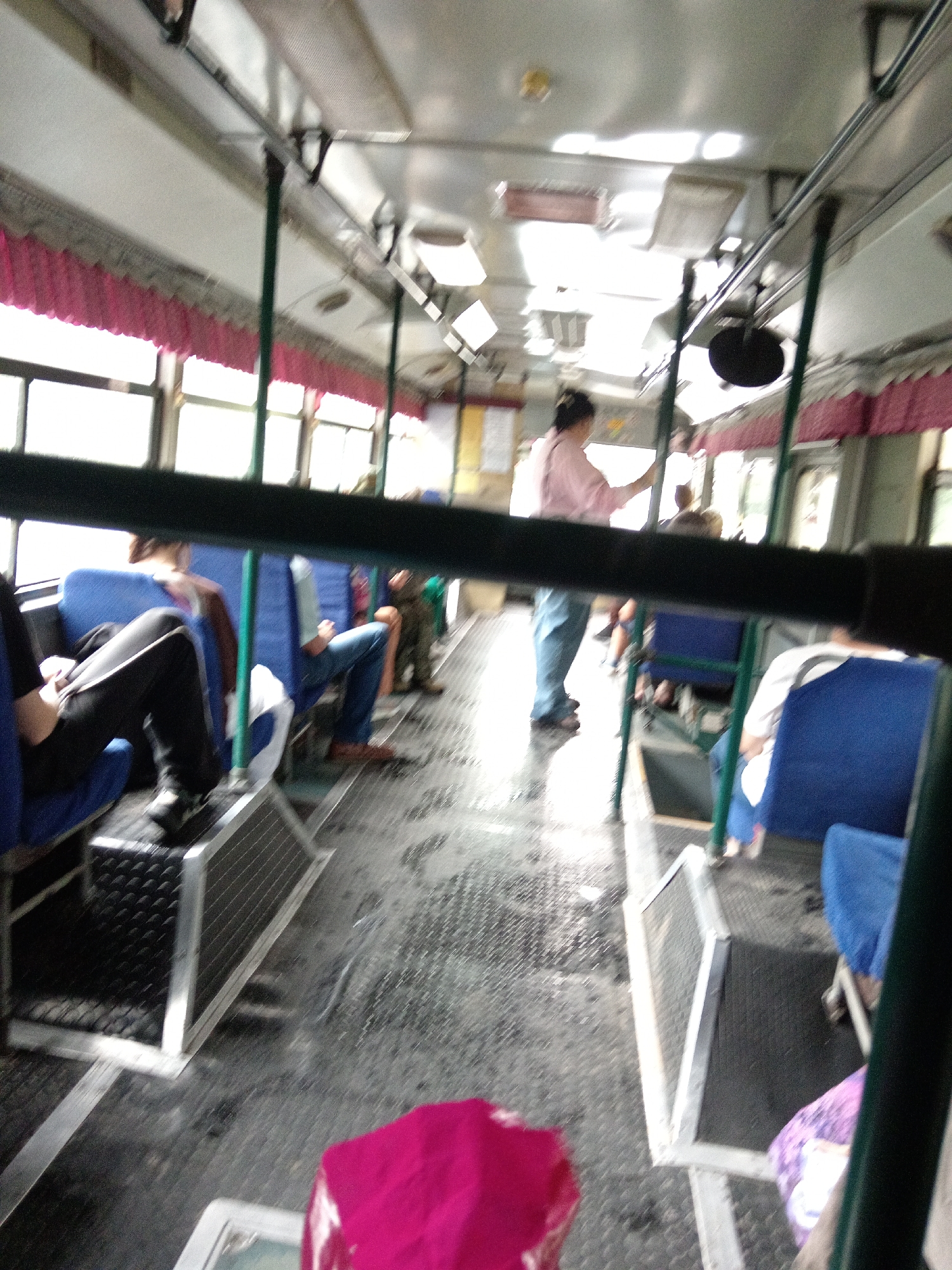 The image depicts the interior of a bus, likely in Belogorsk, Russia, with several passengers seated and standing. The lighting is somewhat dim, with natural light entering from the windows, which are partially obscured by pink curtains.
In the foreground, a person is seated on a blue seat, wearing black pants and a dark top. Their legs are visible. To their left, another person is seated, facing away from the camera, with their legs and feet visible, wearing blue pants and brown footwear.
Further inside the bus, a person wearing a light pink top and blue pants is standing near the front, appearing to be interacting with someone or reaching for something. Another individual is partially visible standing near them. There are other passengers seated in blue seats, some with their backs to the camera.
The floor of the bus appears to be a textured metallic surface, possibly worn or wet in places. Metal poles and railings are visible throughout the interior, supporting the roof and providing handholds for passengers. The overall environment suggests a public transportation setting, possibly during a journey or at a stop.