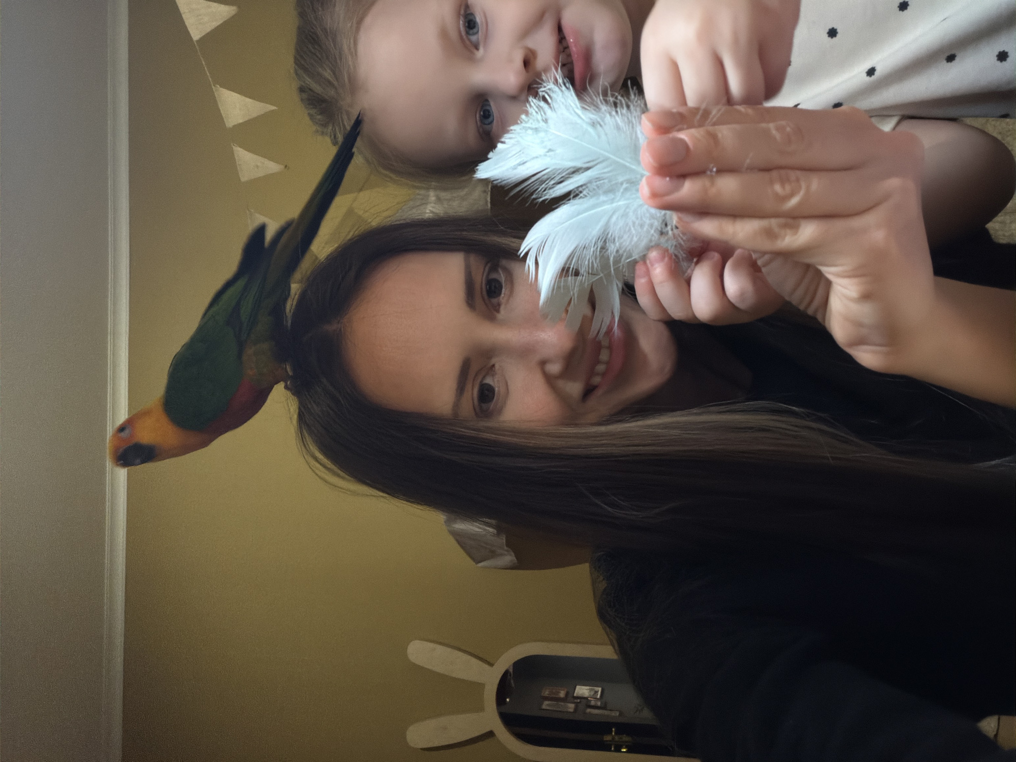 This image depicts an indoor scene in Khimki, Russia, featuring an adult female, a young female child, and a parrot. The adult female, with long, dark hair and wearing a dark garment, is positioned on the left side of the frame, smiling towards the viewer. A multi-colored parrot, exhibiting an orange-yellow head and green body, is perched on her head, facing left. The young female child, with light-colored hair and blue eyes, is partially visible on the right, also smiling towards the viewer. Both individuals are cooperatively holding a cluster of light blue-white fluffy feathers directly towards the camera. The background consists of a plain mustard-yellow wall, surmounted by a white ceiling with a decorative cornice. A string of light-colored triangular pennants is visible against the yellow wall in the upper-right quadrant. To the left, a wooden, rabbit-ear-shaped object, possibly a mirror or decorative frame, reflects an interior room with framed pictures on a dark wall and a visible door handle. The overall environment suggests a domestic setting.
