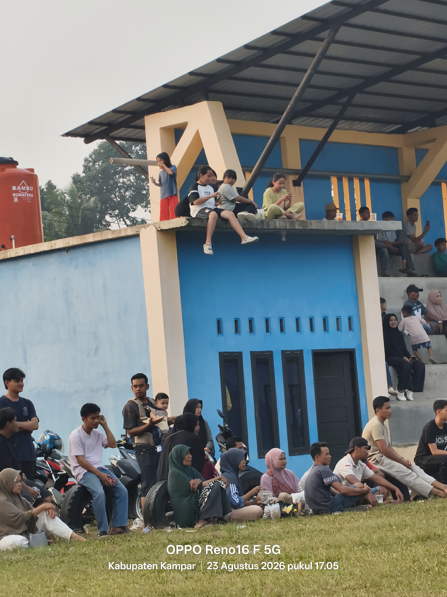 In the city of Simalinyang, Indonesia, a crowd of spectators sits on the grass and concrete bleachers of a sports stadium. A bright orange water tank, branded "BAMBU SUMATERA," stands to the left of the frame. People are gathered both on the ground and on the elevated seating area, some are watching an event, while others are socializing. A man in the foreground is holding a baby in a carrier. The stadium appears to have blue walls and a light-colored roof structure supported by thick poles. The lighting suggests it is late afternoon.