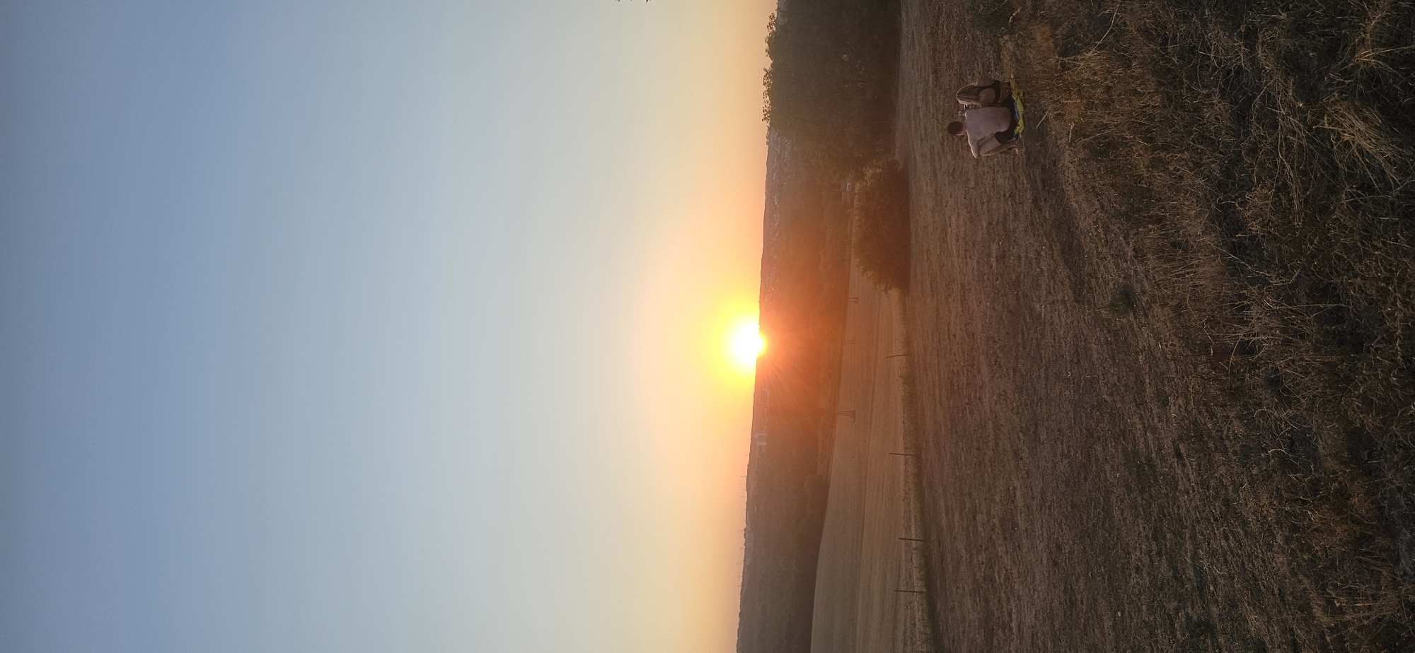 The image captures a serene sunset over a rural landscape, likely on the outskirts of Keszthely, Hungary. The sky transitions from a clear, pale blue at the top to vibrant hues of orange and yellow near the horizon, where the sun is setting. The sun itself is a bright, distinct orb just above a distant tree line and faint city silhouette.
In the foreground, a couple is seated on a yellow blanket on a grassy slope. They are facing away from the camera, looking towards the sunset. The man is shirtless, wearing dark shorts, and the woman is wearing a dark bikini top. They appear relaxed, with the woman leaning slightly against the man.
The terrain is characterized by dry, mown grass and earth, with a few sparse tufts of taller, dried vegetation. A simple wire fence runs horizontally across the middle ground. Beyond the fence, gently rolling hills are visible, dotted with agricultural fields and patches of trees. In the distance, the silhouette of a town or city is discernible against the bright sunset, with some faint structures and possibly wind turbines visible on the hills. The overall atmosphere is peaceful and natural, indicative of an evening spent enjoying the outdoors.