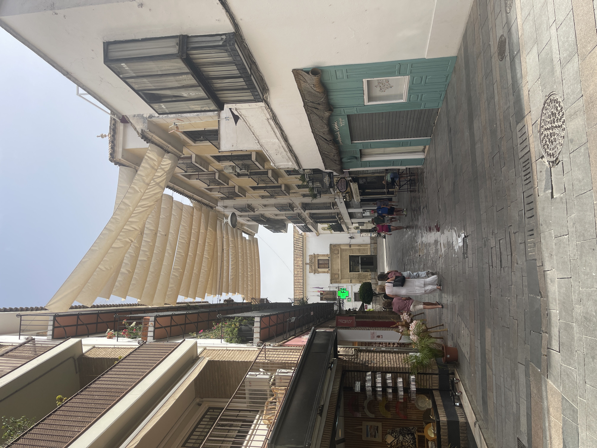The image captures a narrow, paved street in Córdoba, Spain, lined with buildings on both sides. A white and cream awning made of fabric panels stretches across the street, providing shade. On the left, a building features balconies with wrought-iron railings and potted plants. Below the balconies, shops are visible, one displaying decorative fans and other merchandise. On the right, buildings are painted white and yellow, with more balconies and windows. One shop on the right has a teal-colored facade with a straw-thatched awning and closed shutters.
Several people are walking down the street. Three women are in the foreground, walking away from the camera. One woman wears a white dress, another a pink shirt and white shorts, and the third a pink top and white shorts. Further down the street, more individuals are walking, including a man in a blue shirt and dark shorts, and two women in the distance. The cobblestone street appears wet, suggesting recent rain or cleaning. In the background, a large building with an ornate facade and a pediment is visible at the end of the street, possibly a church or public building, with flags on either side of the entrance. A green cross sign with the letters "FB" is mounted on a pole near this building, indicating a pharmacy. The sky is bright and clear, suggesting a sunny day.