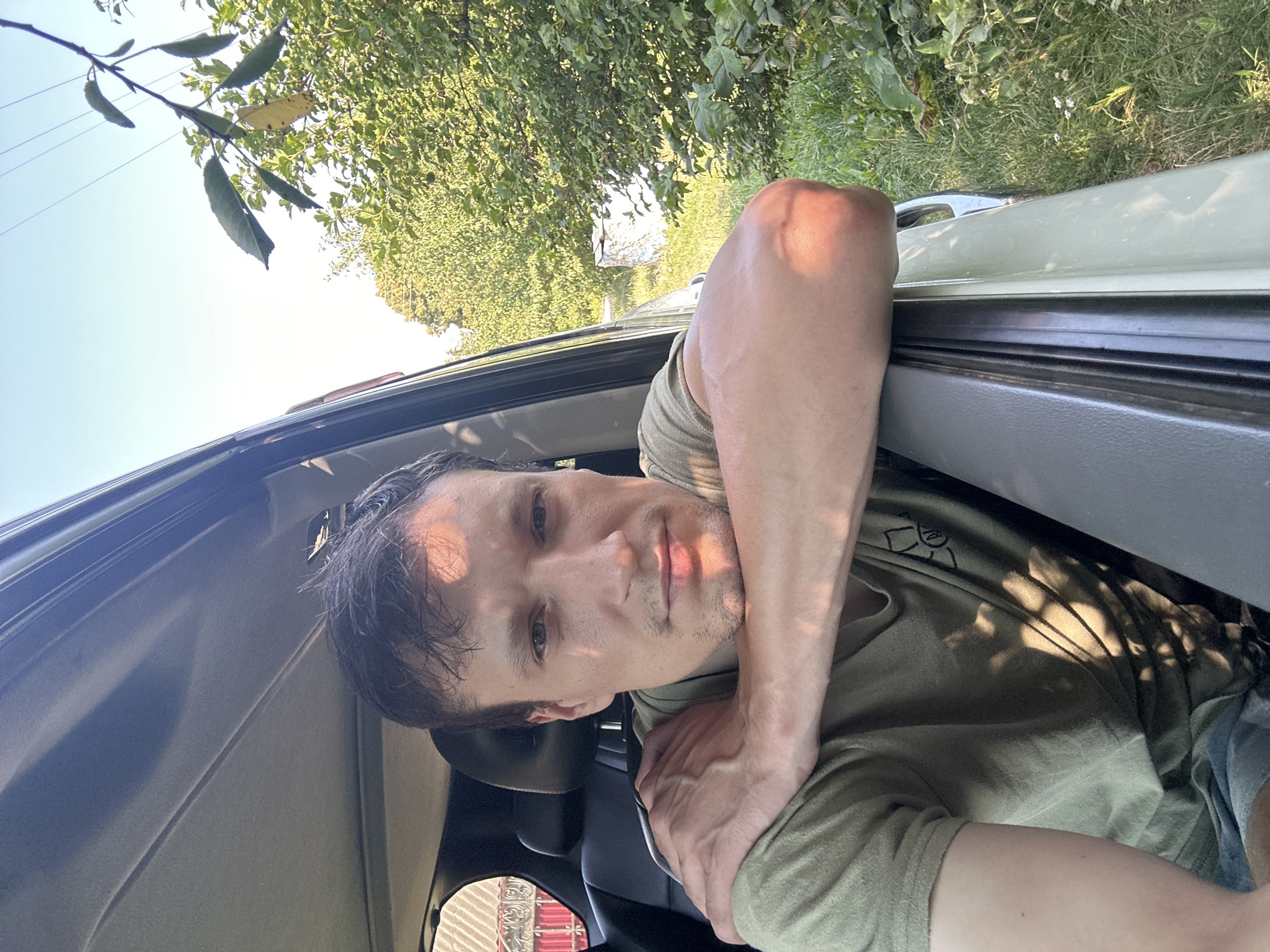 A man with short, dark hair and a light beard is shown from the chest up, leaning his arm out of a car window. He is wearing a green t-shirt and blue shorts. The car appears to be parked on a grassy area with trees and foliage in the background. The lighting suggests it is daytime, with sunlight casting shadows on the man's face and arm. A white cylindrical object and some fencing are visible in the distance among the trees. The overall scene is outdoors, with the car door frame and interior visible in the foreground.