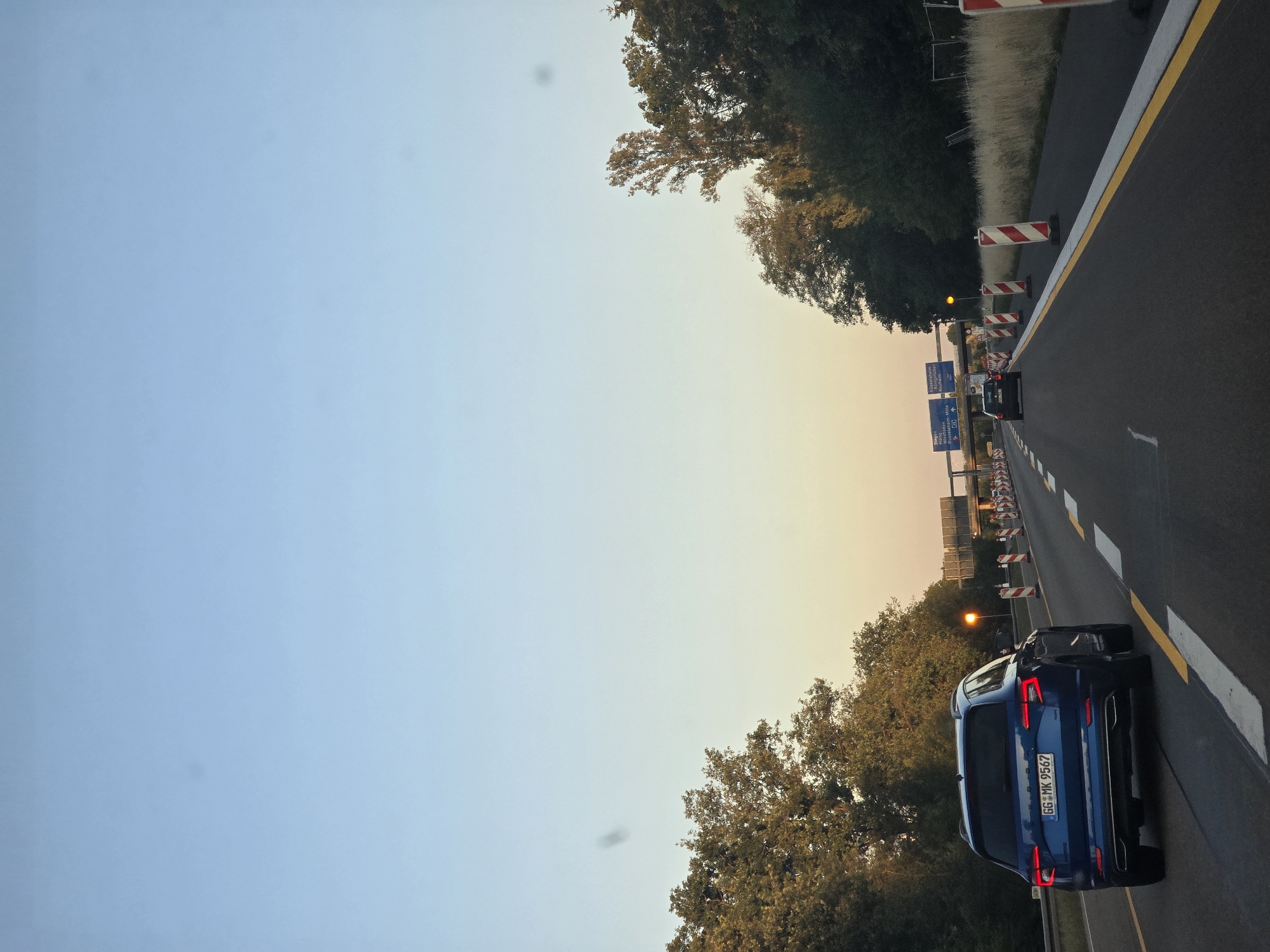 The image shows a blue Skoda Karoq driving on the Autobahn in Rüsselsheim am Main, Germany, during sunset. The car is in the left lane, and the road ahead is undergoing construction, with traffic cones and barriers visible. In the distance, highway signs indicate directions to Rüsselsheim, Mainz, and Frankfurt Airport. Trees line both sides of the road, and the sky is a clear, pale blue transitioning to a warm orange hue near the horizon. There are no people visible other than the driver and any potential passengers inside the vehicles.
