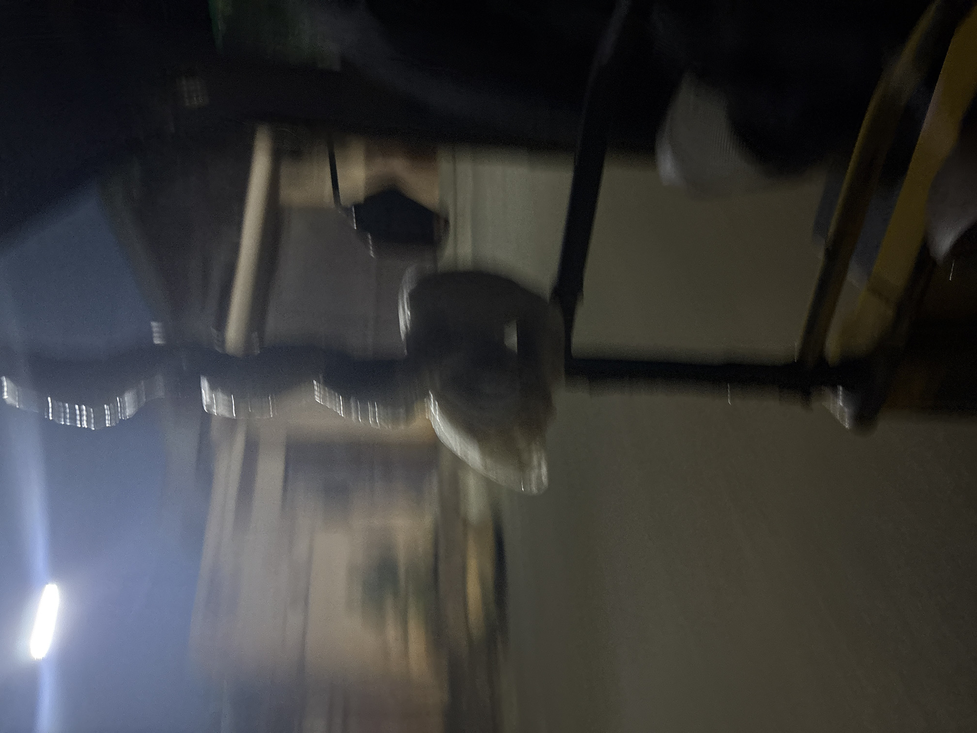 The image depicts a motion-blurred view from the interior of a vehicle, likely a tuk-tuk or similar passenger vehicle, in Onitsha, Nigeria. The perspective is from the passenger area looking outward and slightly upward. In the foreground, a dangling object, possibly a bag or bundle wrapped in transparent plastic, is suspended from a metal bar. The object is indistinct due to motion blur but appears to have a light-colored filling.
To the left of the suspended object, a blurred background shows a street scene at night. A bright, elongated light source, possibly a street lamp or sign, illuminates the upper left portion of the frame. Adjacent to this, indistinct buildings or structures with muted colors are visible, suggesting an urban environment. The street itself appears to be paved and dark.
To the right of the suspended object, the interior of the vehicle is visible, primarily in shadow. Parts of the vehicle's structure, including a dark metal pole and what appears to be a yellow upholstered seat or component, are present. The overall impression is one of movement through the city at night, with the primary focus on the dangling object within the vehicle. The motion blur conveys a sense of travel.