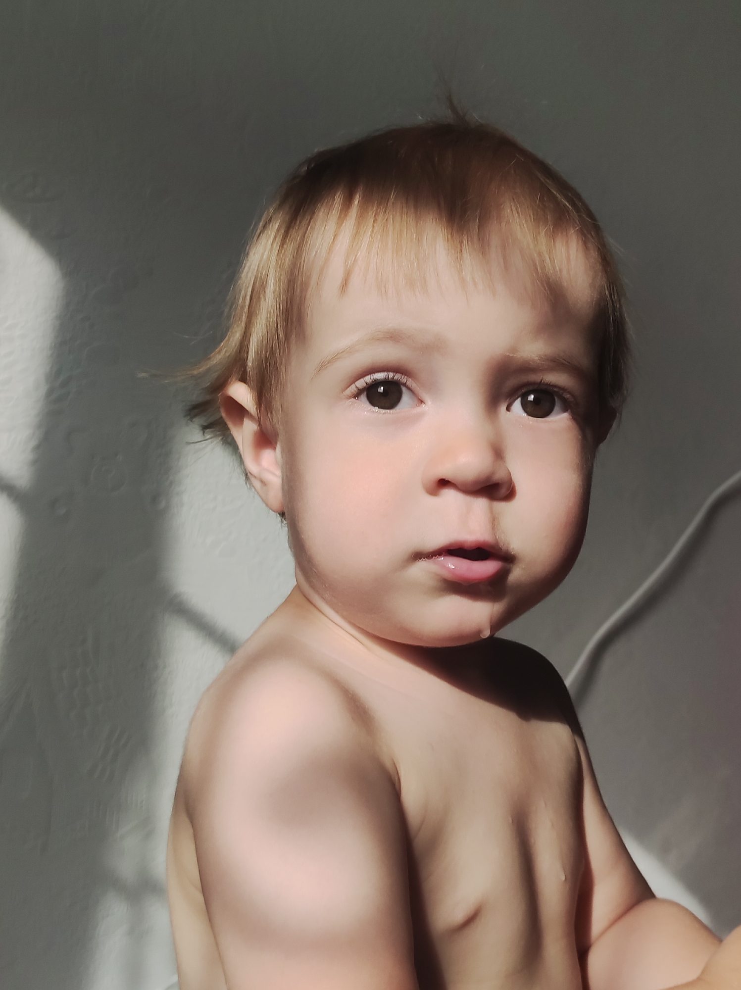 A toddler with fair skin, blonde hair, and brown eyes is centered in the frame, looking directly at the camera. A small drop of liquid is visible at the corner of their mouth, suggesting drool. The child's upper torso is bare. The background is a plain, textured wall, partially illuminated by natural light, casting a shadow to the left of the child. A white electrical cord is visible running vertically along the wall in the background, to the right of the child. There are no overt indicators of location within the city of Chaikovsky, Russia.