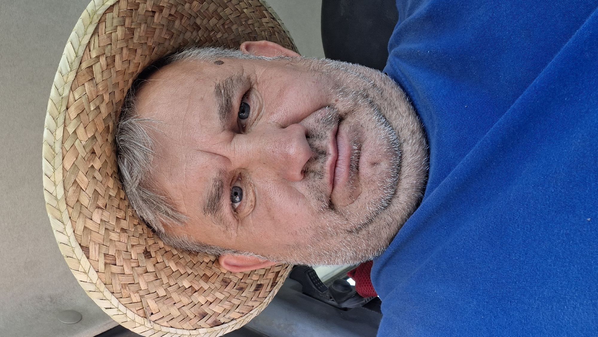 A close-up, selfie-style photo shows a middle-aged Caucasian man wearing a straw hat and a blue t-shirt. He has grey hair, a grey beard, and blue eyes. The man is looking directly at the camera with a neutral expression. The background is blurred, but appears to be the interior of a vehicle, possibly a car or a tractor. There is a red object visible behind the man's left shoulder, and a grey plastic surface behind his hat. The image is well-lit, suggesting it was taken during the day. The location is not discernible from the image itself, and there is no information to indicate it is in Hîncești, Moldova.