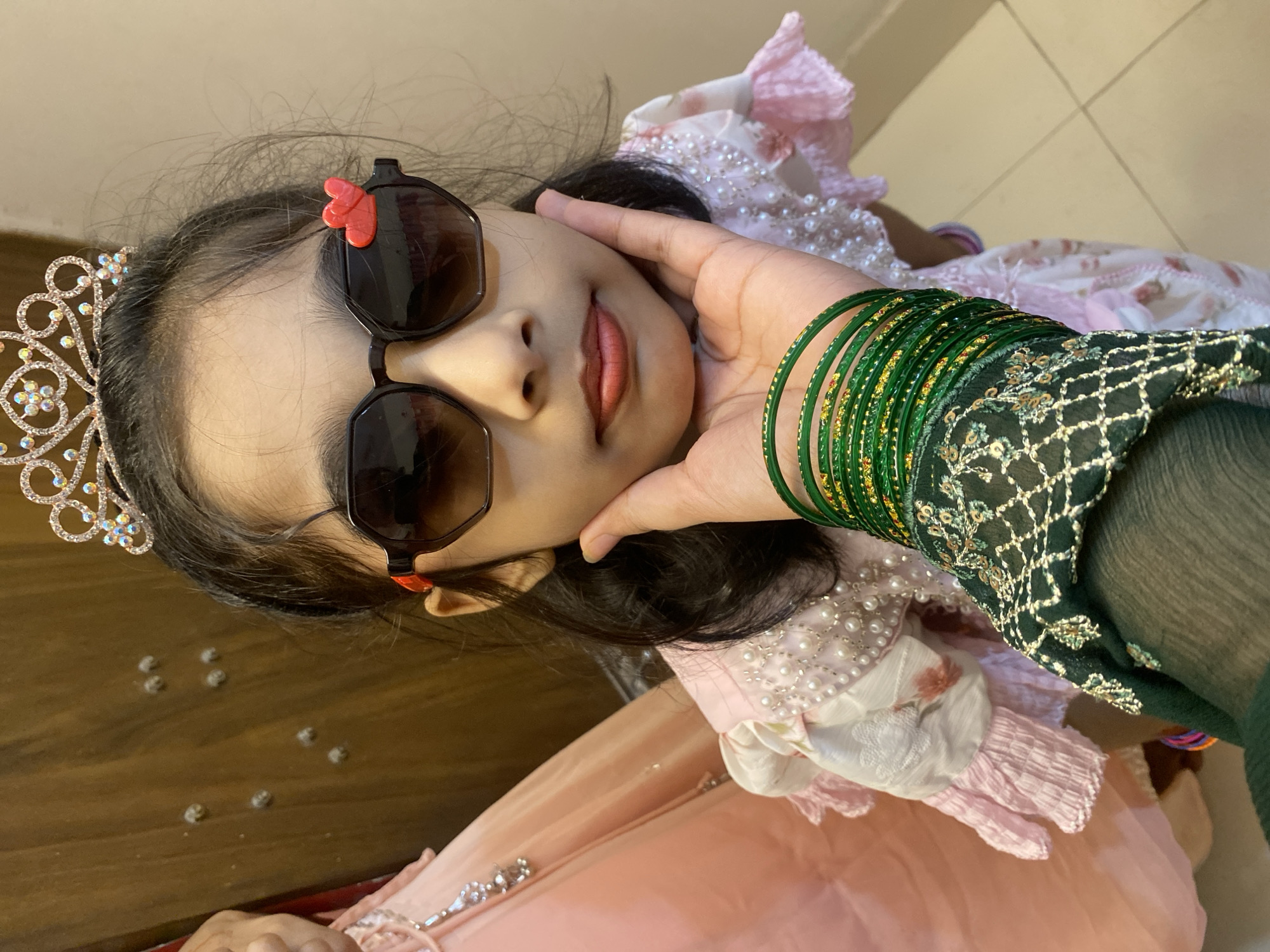The image shows a close-up of a young girl, likely in Dhaka, Bangladesh. She is wearing a sparkly tiara, dark sunglasses with a small red heart embellishment on the frame, and a pink dress adorned with pearls. A woman's hand, adorned with numerous green bangles, gently supports the girl's cheek. The background is blurred, but appears to be an indoor setting with wooden surfaces and tiled flooring. The lighting suggests it could be daytime, possibly indoors or in a shaded area. The girl's expression is relaxed and content. The overall impression is one of warmth and affection.