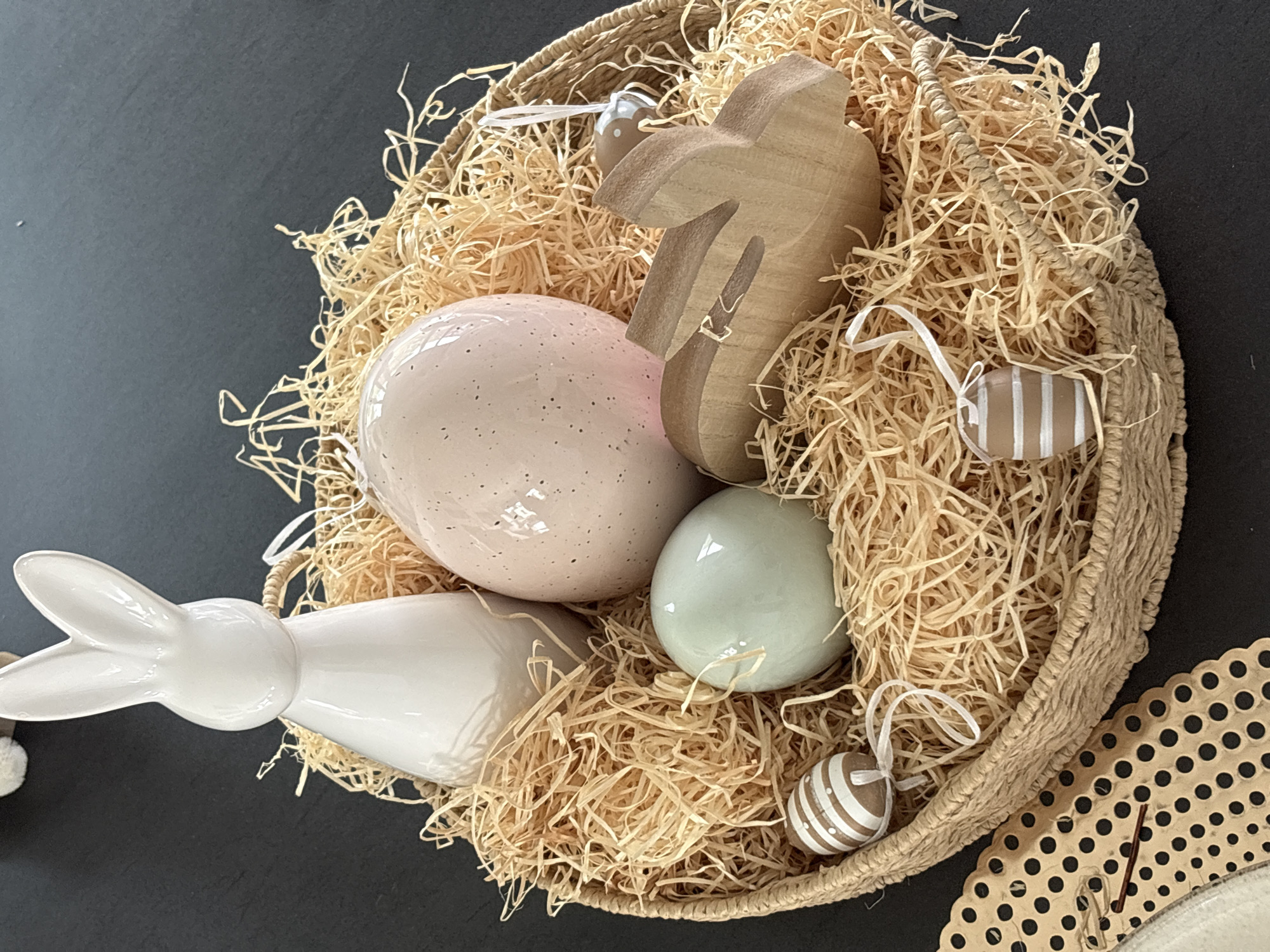 A high-angle, close-up shot depicts an Easter basket situated in Junglinster, Luxembourg. The basket, woven from light brown natural fibers, is filled with tan-colored straw-like material resembling wood shavings. Within this nest are various Easter decorations: a large, pale pink speckled egg; a light green egg; a white ceramic bunny; a wooden bunny silhouette; and two small, brown eggs with white stripe decorations, each suspended by white ribbons. The basket is set against a dark, matte surface. To the lower right corner, a portion of a light tan, perforated material with dark circles is visible.