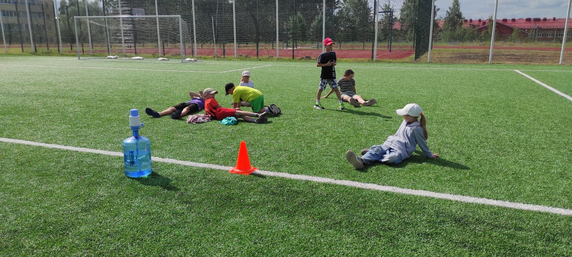 The image shows a group of children on a bright green, artificial turf soccer field in Noviy, Russia. In the foreground to the left, a large blue water cooler bottle with a white and blue pump is visible. Several children are scattered across the field. Three children are lying down near the water cooler, with one in a purple shirt and black shorts, another in a red shirt and black shorts wearing a beige cap, and a third in a neon green shirt and green shorts. A fourth child, a girl wearing a white cap and grey hoodie, sits facing right on the grass. Further in the background, a boy in a black t-shirt and camouflage shorts stands facing left, and another child in a striped shirt sits on the grass. An orange traffic cone is placed on a white line marking the field. In the background, a soccer goal, fences, trees, and buildings with red roofs are visible. The sky is partly cloudy.