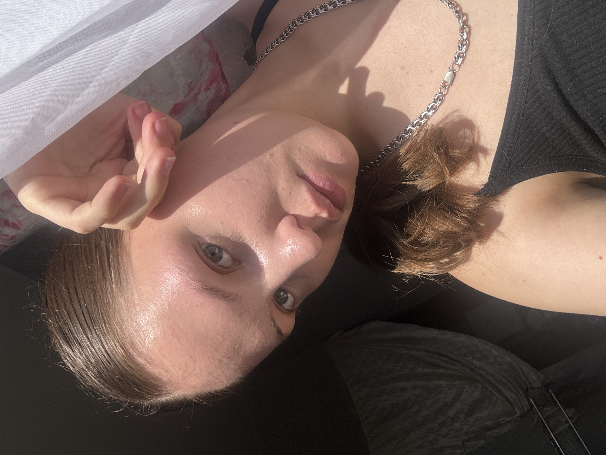 The image shows a close-up of a young woman's face and upper body. She has fair skin with some visible pores and a slight sheen, possibly from sweat or skincare. Her hair is pulled back and appears wet or slicked. Her eyes are green and her lips are slightly parted. She is wearing a black tank top and a thick, silver chain necklace. Her right hand is raised to her face, with her fingers touching her temple. The lighting suggests a bright, sunny day, with strong shadows cast across her face and body. A white, sheer curtain is visible in the upper right of the frame. The background is dark and out of focus, with some texture that could be a blanket or upholstery. There is no identifiable information within the image that can be used to determine its location in the city of Марковский, Russia.