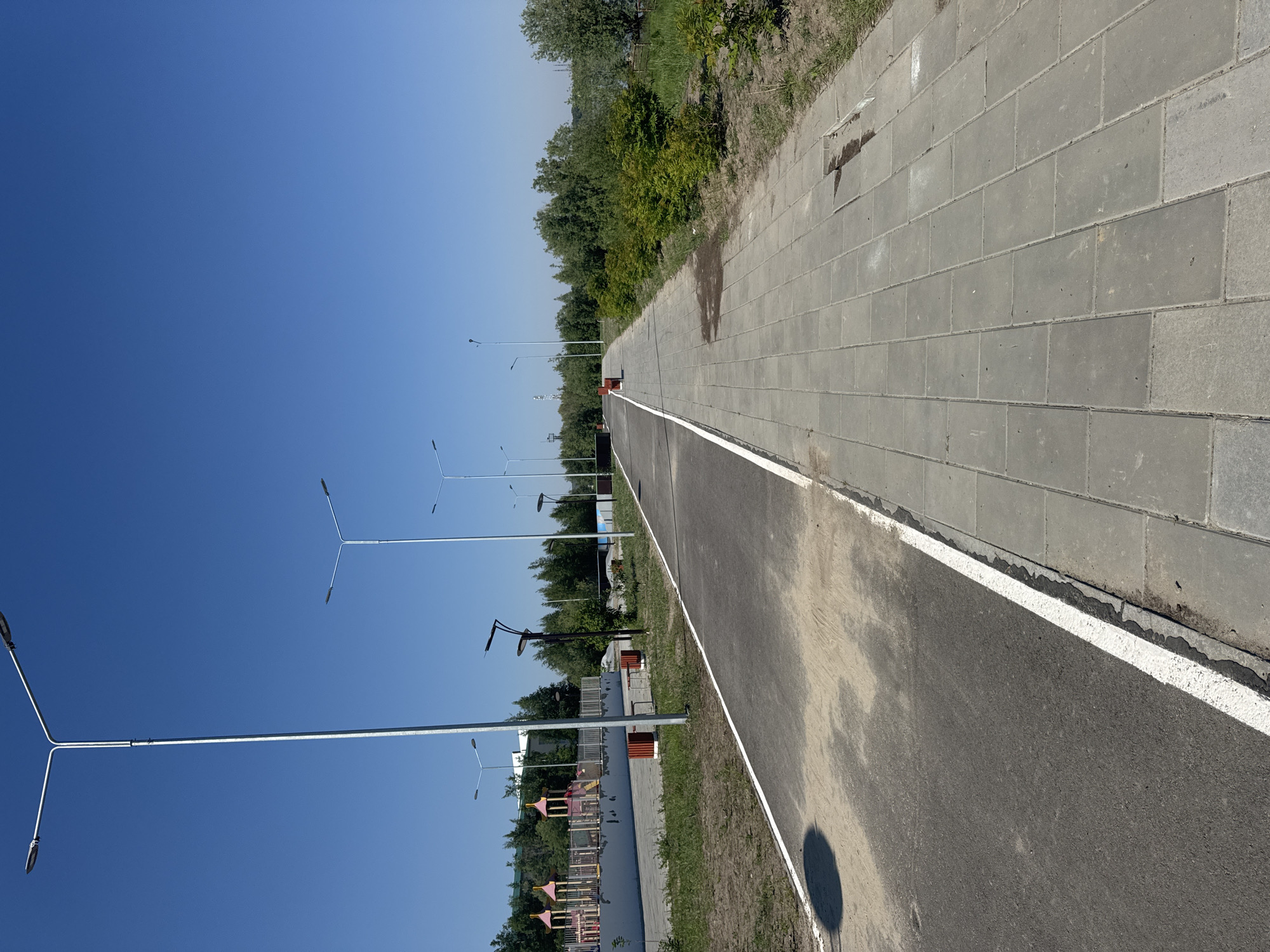 This image shows a paved pathway alongside a playground and a body of water in Megion, Russia. The pathway is divided into two sections: a darker asphalt section on the left, possibly a bicycle or rollerblade path, with a white line marking its edge, and a lighter gray paved section on the right, suitable for pedestrians. Several tall lampposts are visible along the pathway.
To the left of the pathway, there is a playground with colorful structures, surrounded by a fence. In front of the playground, there are some red benches and planters. Further to the left, there are trees and buildings in the background.
The right side of the pathway features green bushes and grass, with a body of water visible in the distance. The sky is clear and blue, indicating a sunny day. The overall scene depicts an outdoor public space designed for recreation and leisure.