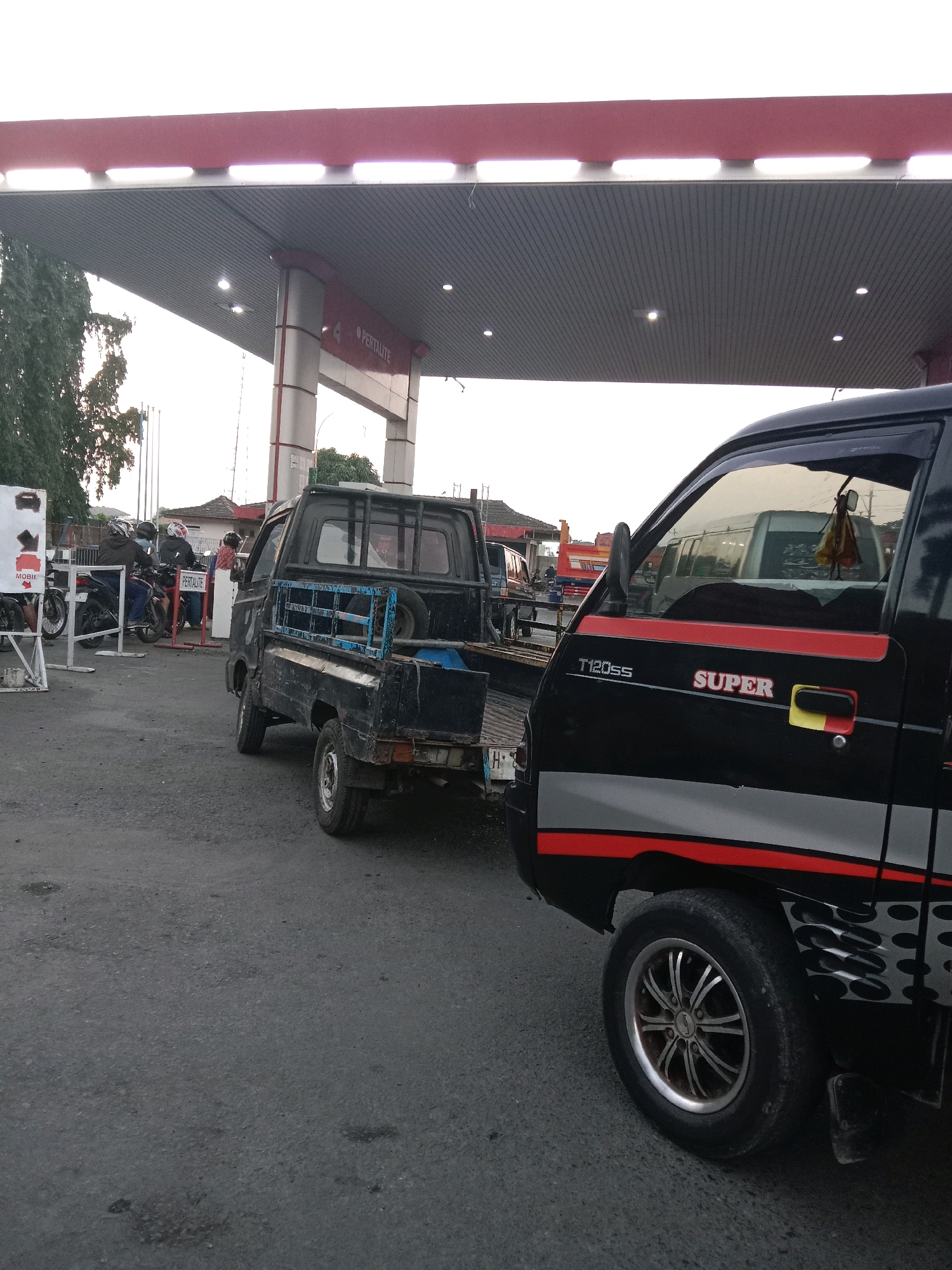 This is a medium-shot, eye-level, outdoor, daylight photograph taken at a gas station in Brangsong, Indonesia. The foreground is dominated by the right side of a black pickup truck with red and grey stripes and silver rims. The truck has a "T120ss" and "SUPER" logo. To the left of this truck is the rear of another black pickup truck, which appears to be towing something. This truck has a blue metal frame around its bed. Further left, there are several motorcycles and people. One person on a motorcycle is wearing a white helmet with red detailing. A white sign with red and black graphics is visible behind the motorcycles. In the background, a gas station canopy with red trim and fluorescent lighting is prominent. The sky is overcast. The ground is asphalt with some dark patches.