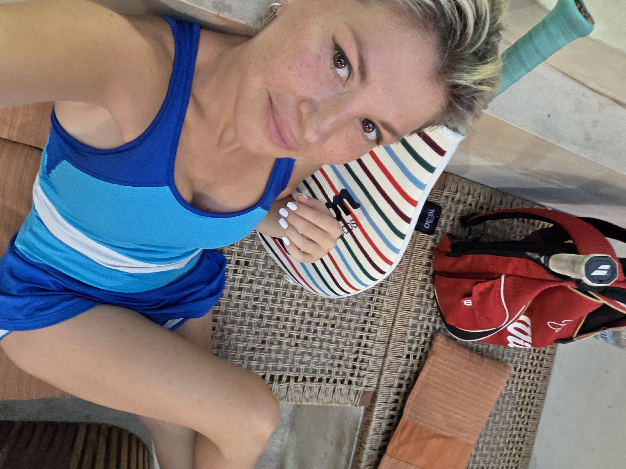The image shows a woman lying down, looking at the camera. She has blonde hair and is wearing a blue athletic outfit. Next to her is a red backpack and a tennis racket. The scene appears to be outdoors on a woven mat, possibly in a resort or recreational area. The woman is resting her head on a striped pillow. The lighting suggests it is daytime.