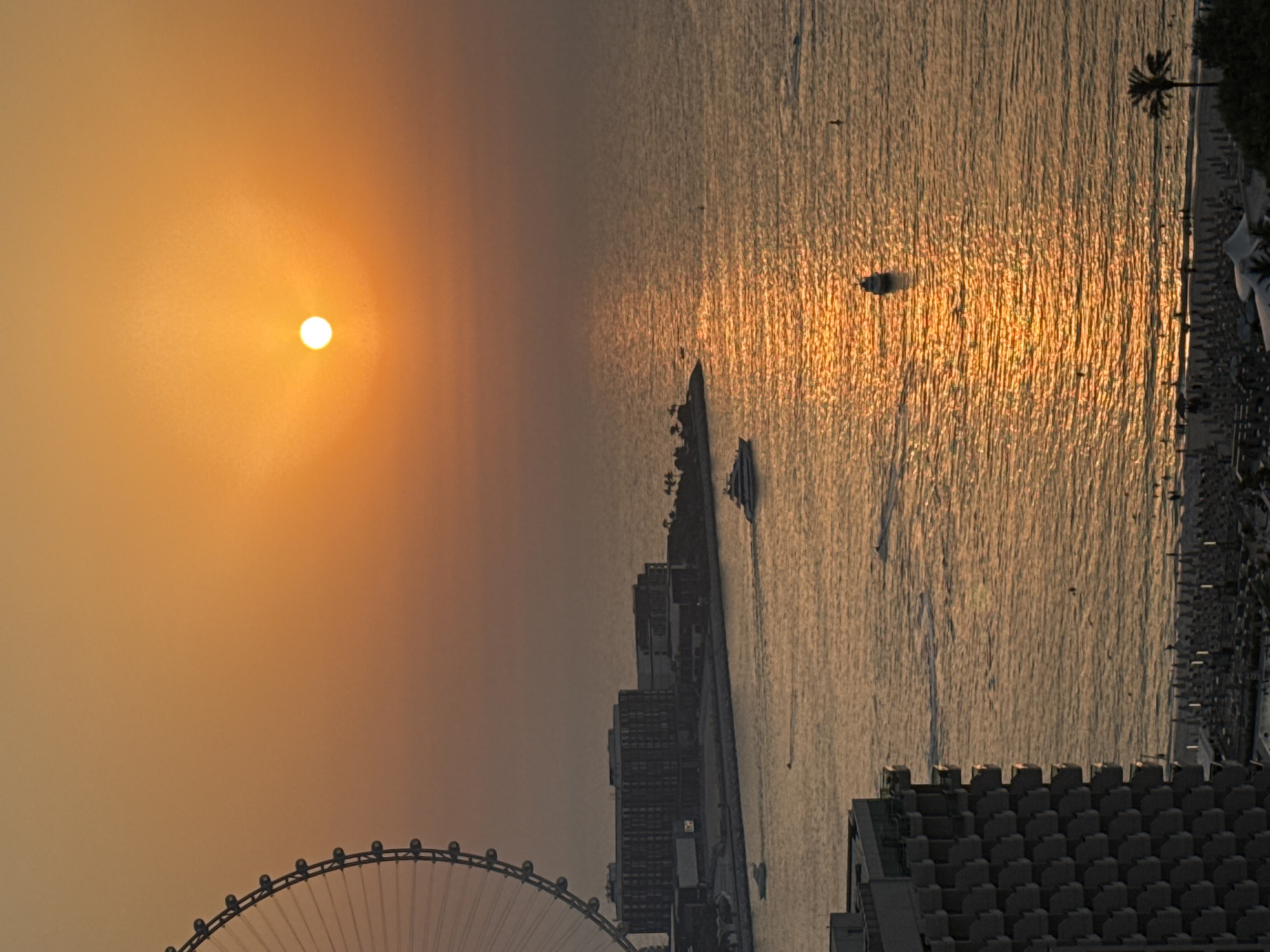 An aerial view captures a sunset over the Arabian Gulf in Dubai, United Arab Emirates. The sun, a bright orb, is positioned in the upper right quadrant of the frame, casting a warm, orange glow across the sky and reflecting on the water. A Ferris wheel, partially visible on the left edge of the image, dominates the upper left corner. In the middle ground, a line of buildings, likely hotels or residential complexes, runs along the coastline. Several boats are scattered across the water, including a large yacht and smaller speedboats. The foreground shows a beach with numerous umbrellas and a few people, indicating a resort area. The overall scene is one of tranquility and scenic beauty at dusk.