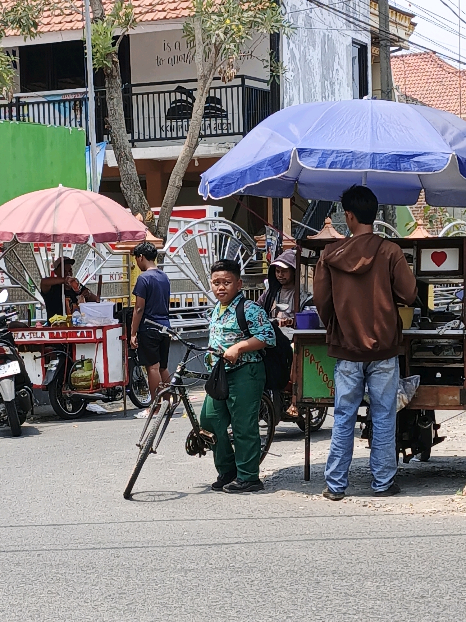 The image captures a street scene in Ketegan, Indonesia, likely in the daytime given the bright sunlight. In the foreground, a young boy wearing a teal and black patterned shirt and green pants stands to the right of the frame, looking towards the viewer with a bicycle in front of him. To his left, a food cart is visible, with a vendor in a dark shirt preparing food under a pink umbrella. Another man in a dark blue shirt and black shorts stands near the cart, looking at the vendor. Further to the right, another food cart is situated under a large blue umbrella. A man in a brown hooded sweatshirt and blue jeans stands with his back to the camera, facing the blue-umbrellaed cart. A red heart graphic is displayed on the side of this cart. The background shows a building with a tiled roof and what appears to be a balcony with a railing. Several motorcycles are parked along the side of the road. The overall impression is a busy street food vendor area.